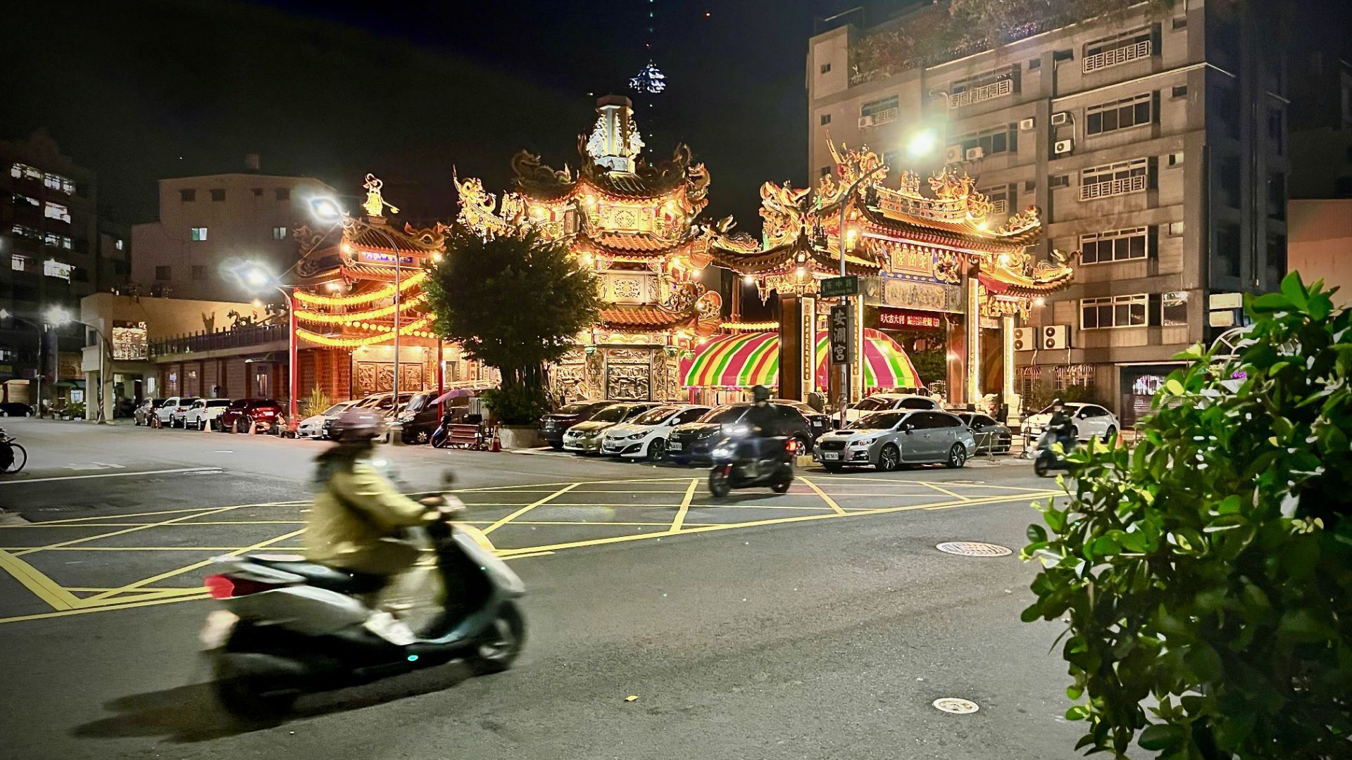 A beautiful red and gold temple at an intersection with scooters riding past.