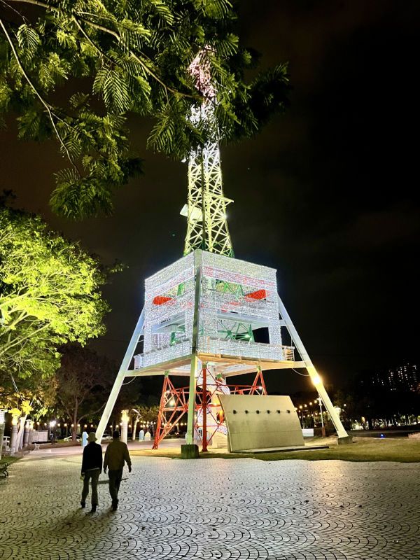 A closer view of the illuminated pylon, with two people walking past. It has an illuminated cube around its center, and a spiral staircase at the bottom.