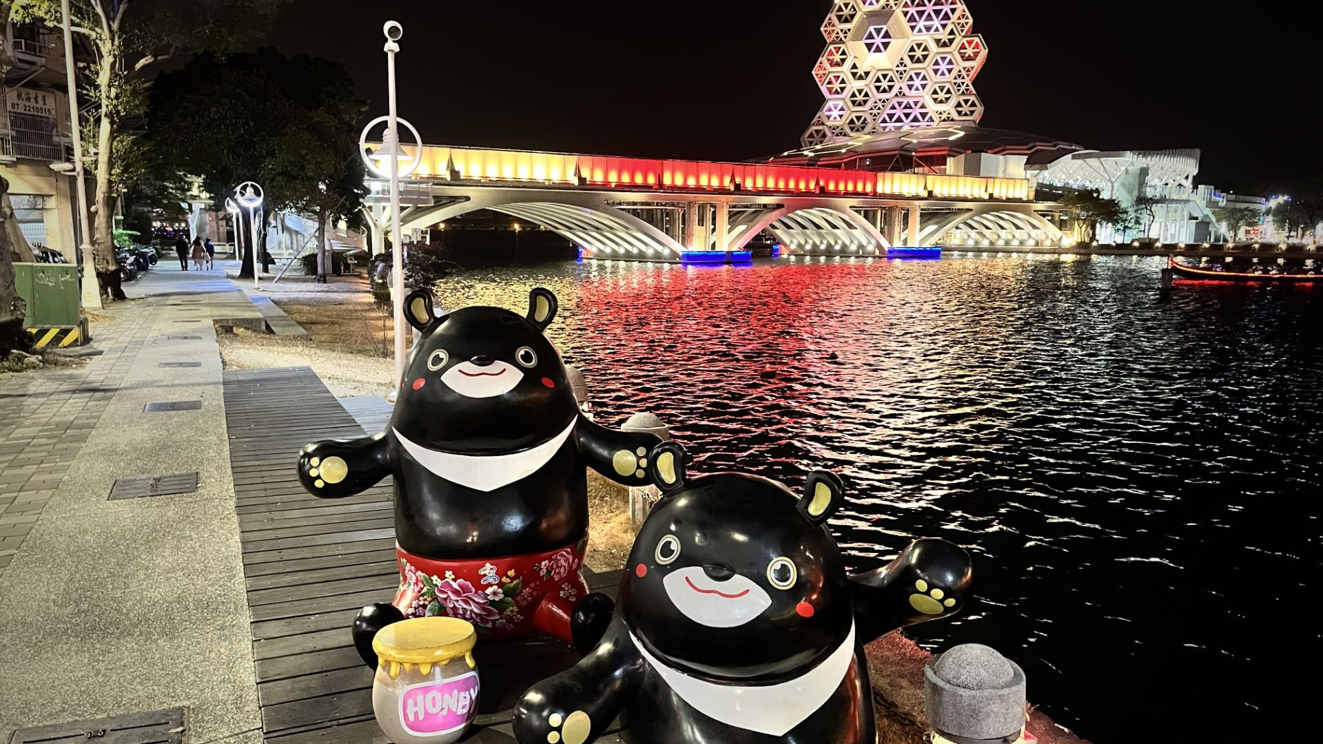 Two Formosan Black Bear statues next to the river, with a bridge and the Kaohsiung Music Center in the background, and an electric gondola gliding by.