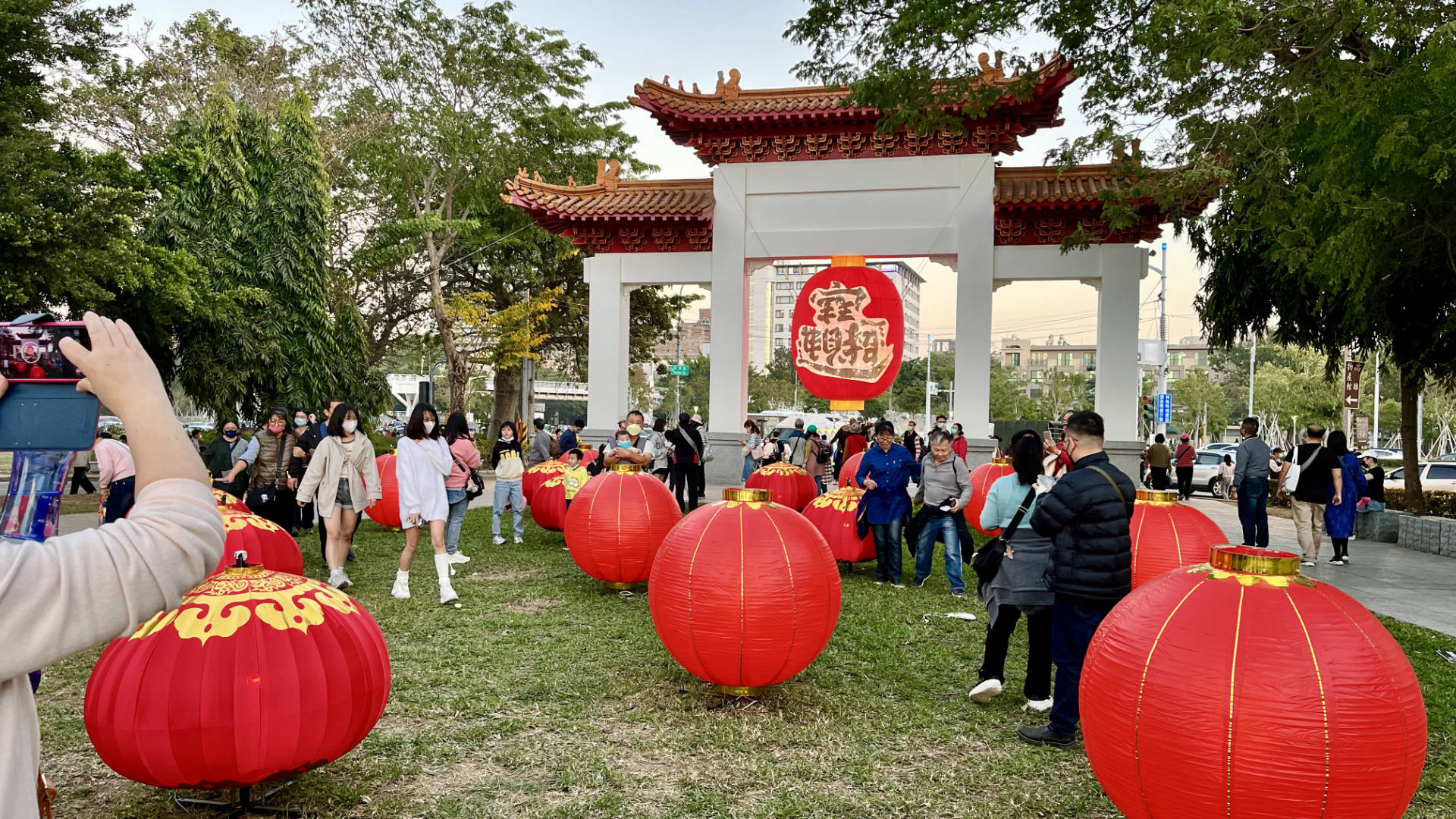 A large lantern hanging from a traditional Chinese gate, with dozens of people taking photos and walking nearby.