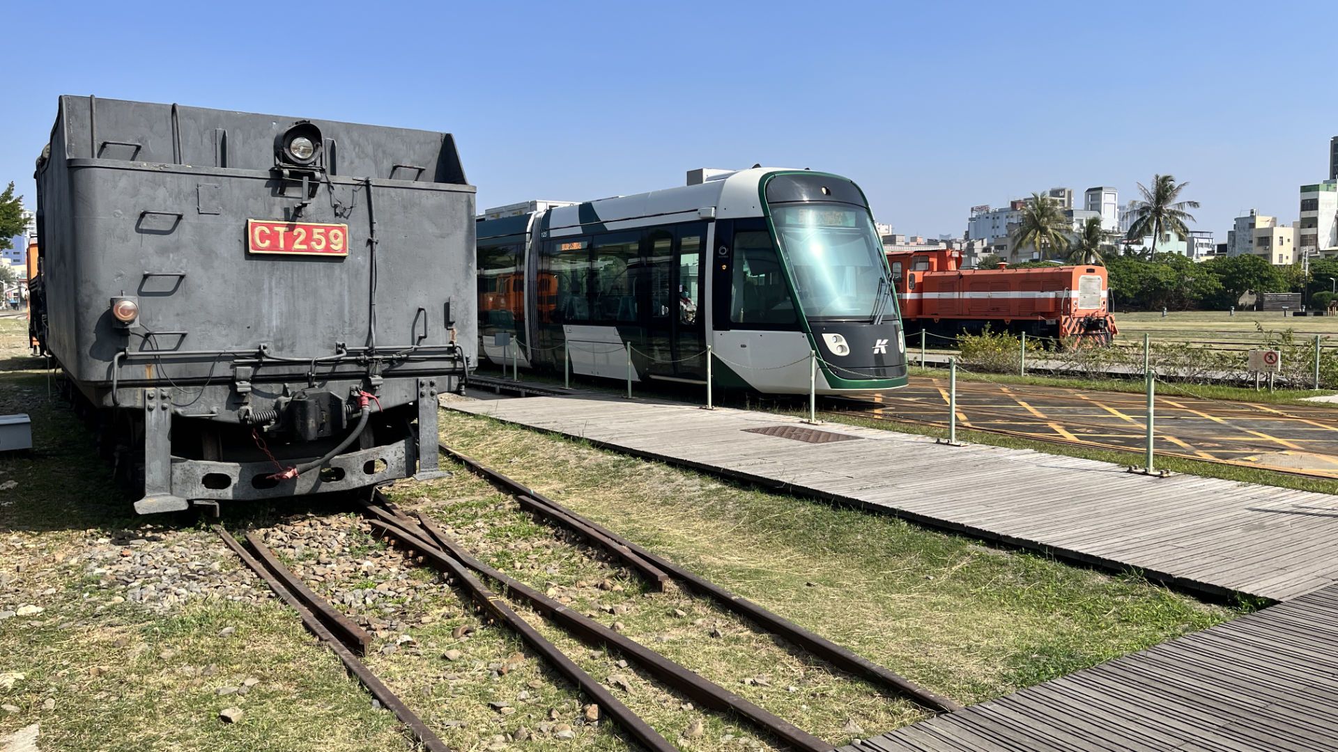 The Kaohsiung LRT tram driving past two stationary historic locomotives, one on either side.