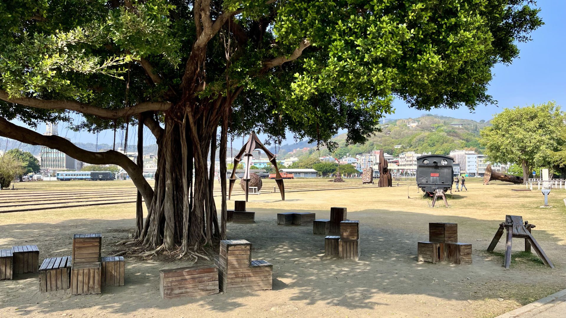 Wooden crate seating under a large tree on the edge of the park, with a historic train and various metal sculptures in the background.