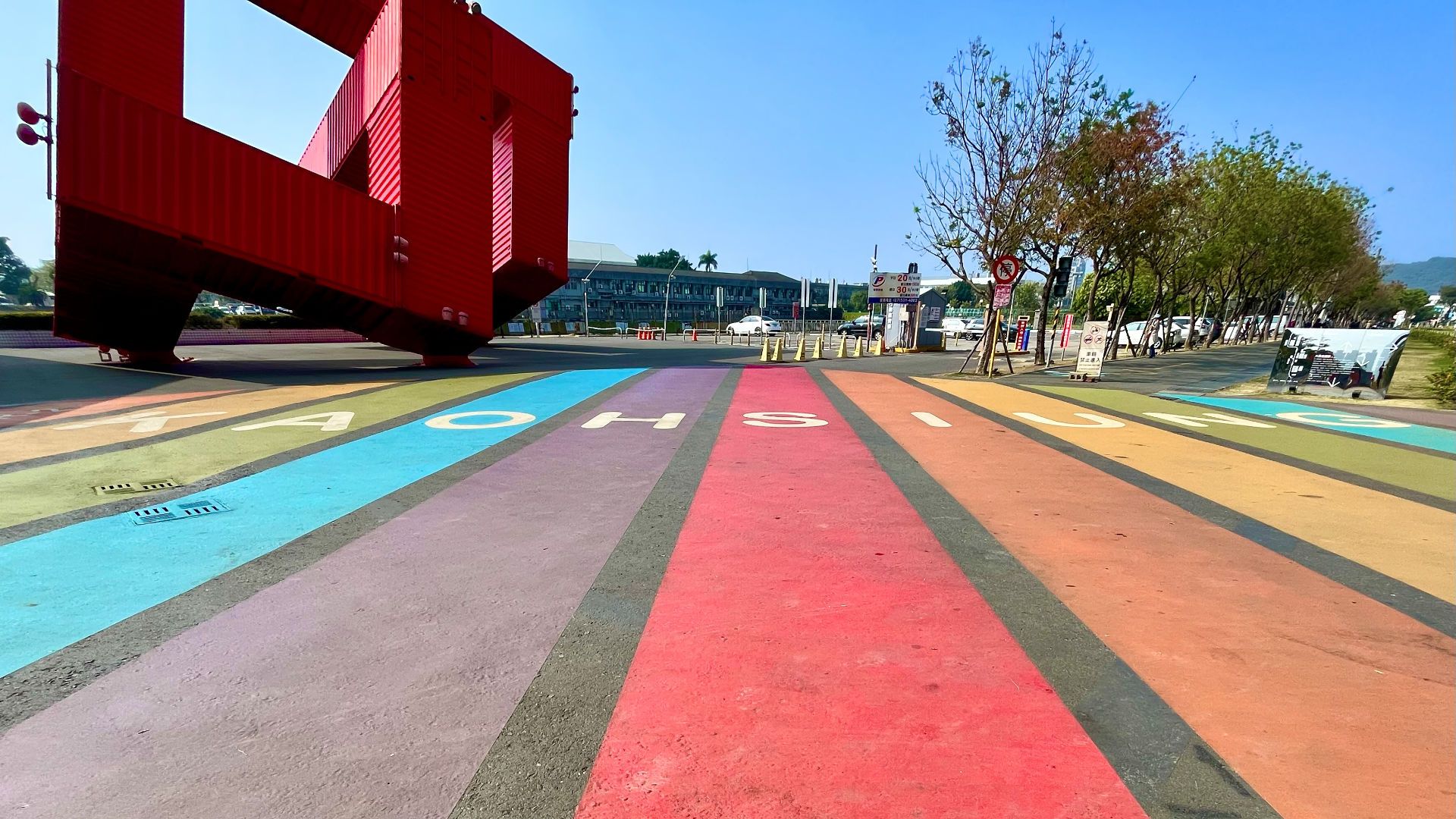 A rainbow crossing with the word ‘Kaohsiung’ written on it, and a large sculpture made of shipping containers in the background.