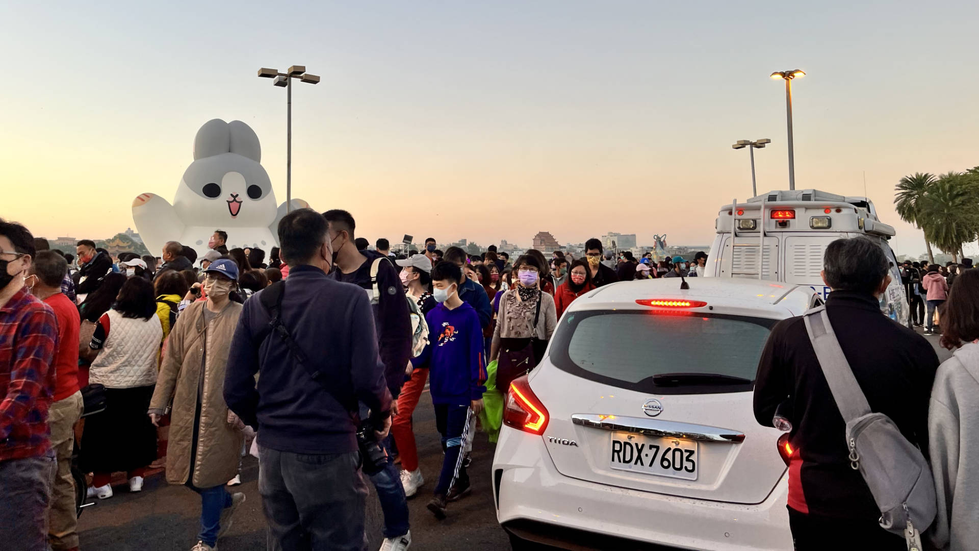 A dense crowd in front of Lotus Point, where an inflatable rabbit is visible in the background. Two vehicles, including a TV news van, are driving through the crowd.