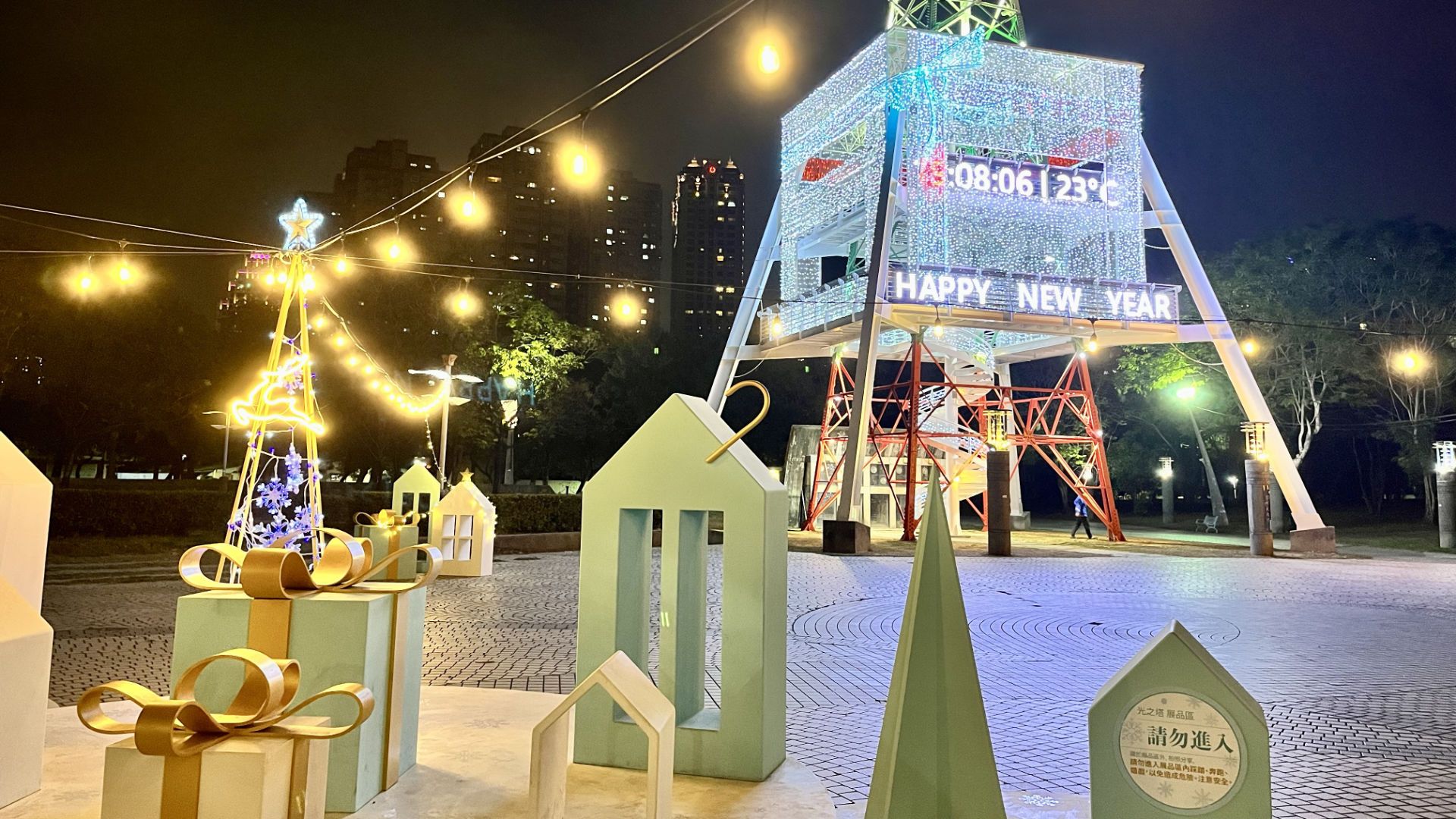 Sculptures of presents and decorations in the foreground, and the current time, temperature, and the words “Happy New Year” illuminated on the side of the pylon.