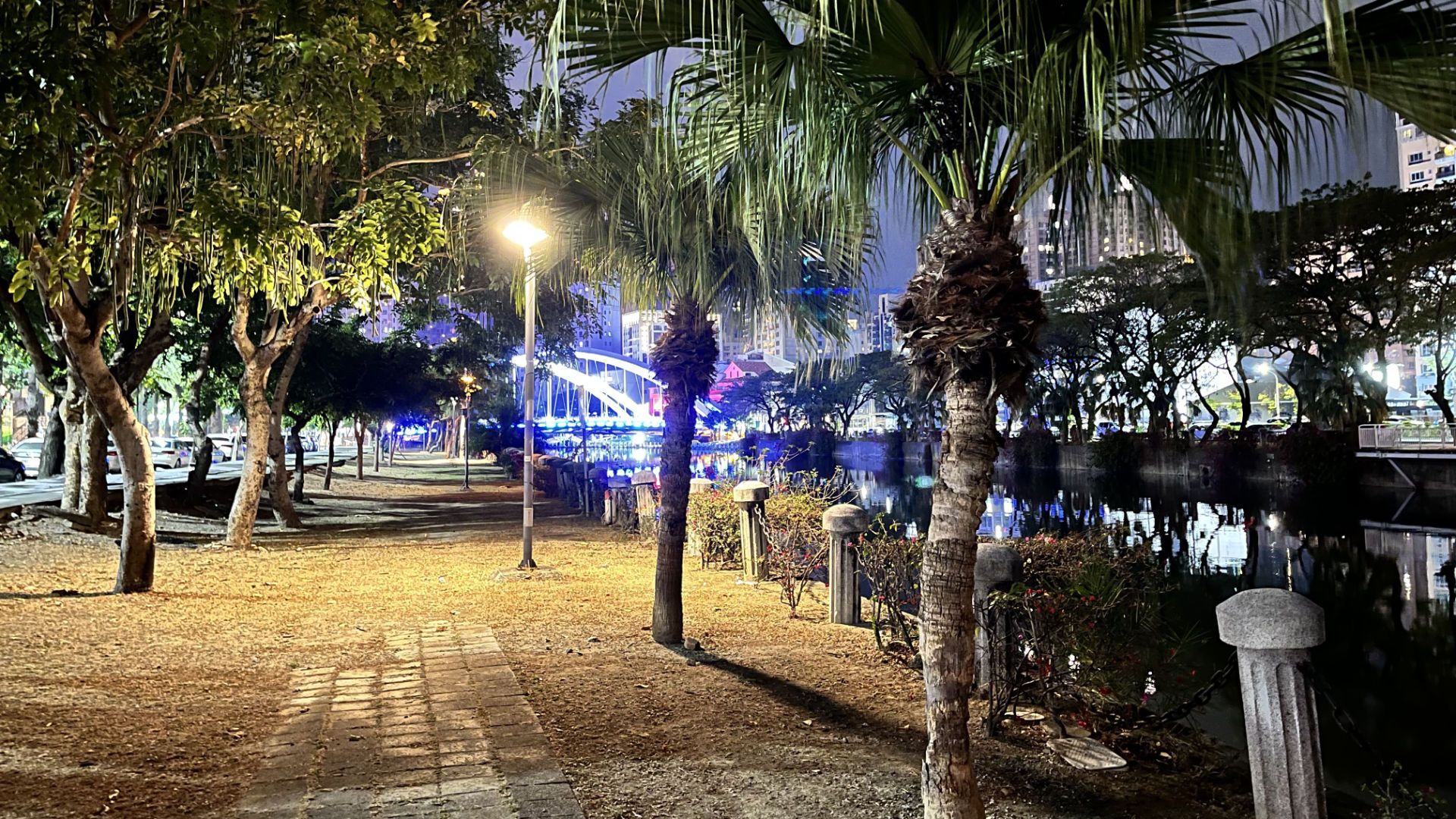 Palm trees line a path next to Love River. An arched road bridge is illuminated in neon blue, in the distance.
