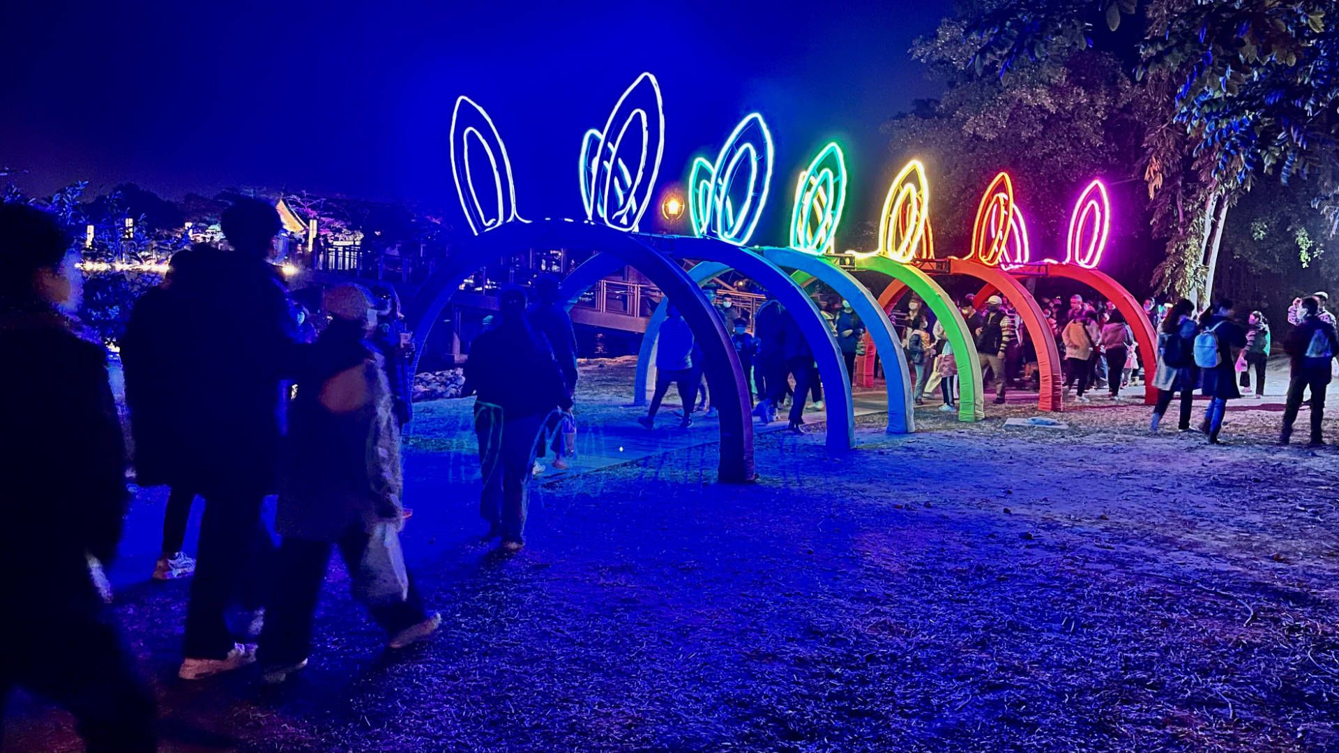 People walking through a sculpture of 7 sets of rabbit ears, illuminated in the colors of the rainbow.