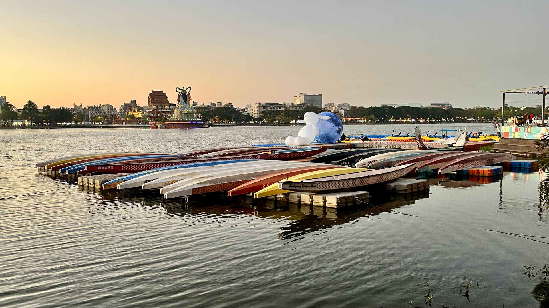 An inflatable rabbit propped against an inflatable moon, on pontoons surrounded by kayaks and dragon boats, on the edge of Lotus Pond.