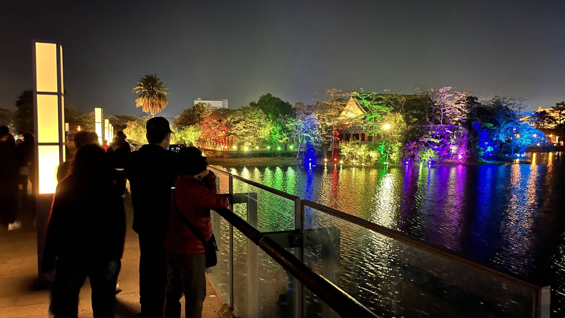 People stand on a bridge looking at a temple across the lake. The temple is surrounded by trees illuminated in rainbow colors.