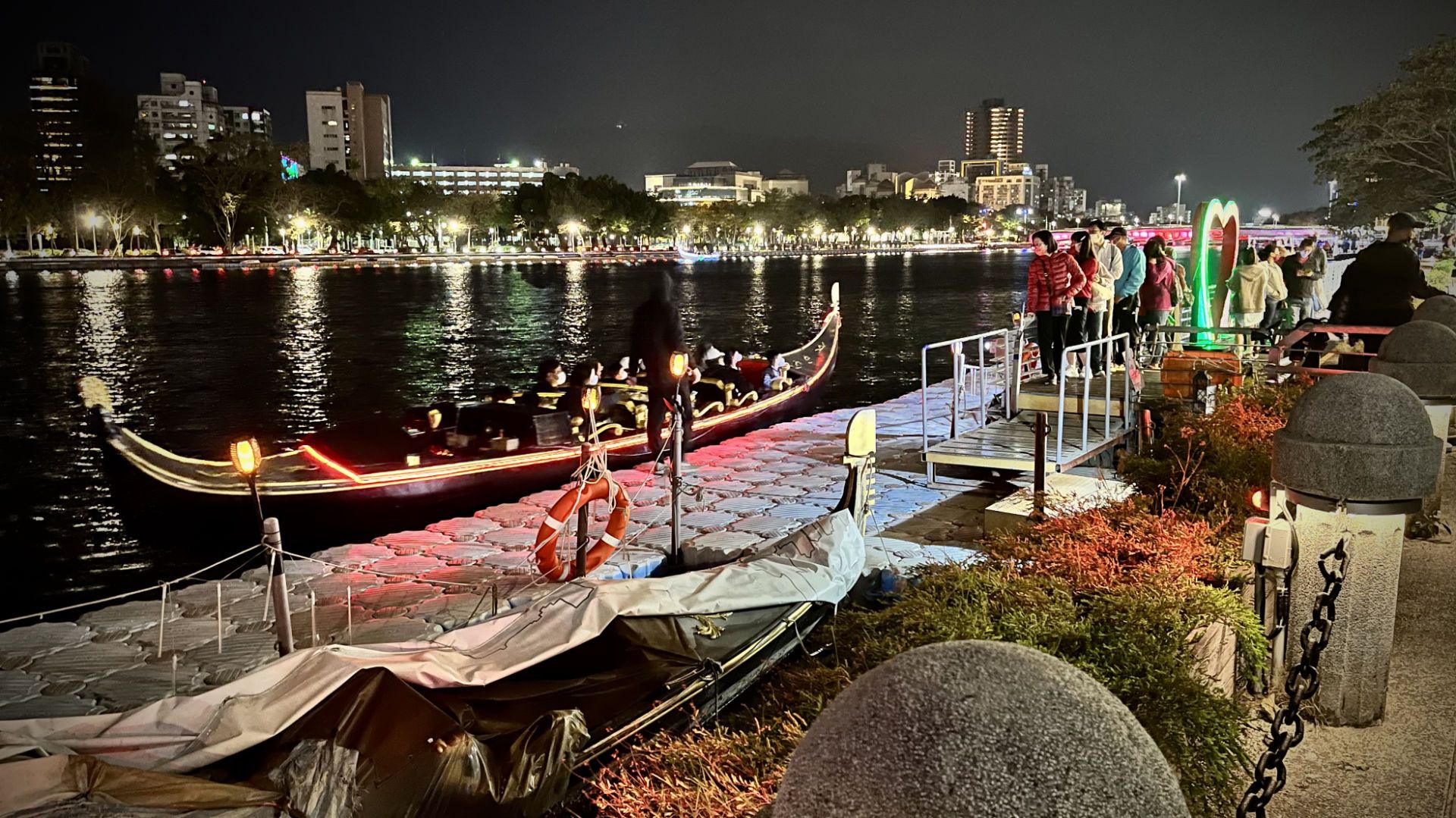 Passengers lined up to board an electric gondola which is docked on the edge of the river. A 2-meter tall heart sculpture is next to them.