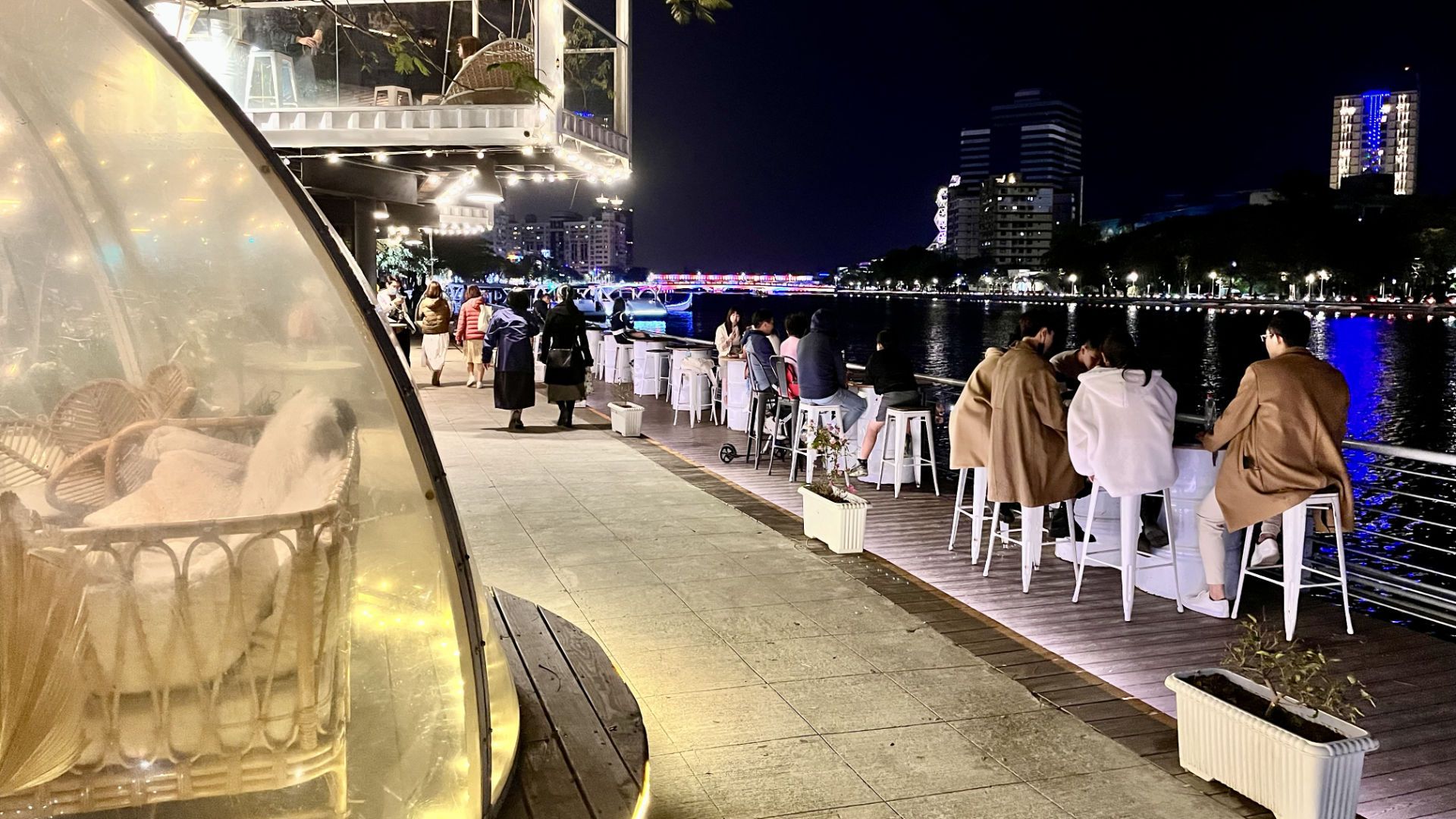 Diners at a bar next to Love River. In the foreground is a spherical room made of transparent plastic, with tropical furniture inside. Above the path is a glass and metal cube-shaped room. Others are sitting on barstools outside, next to the river.