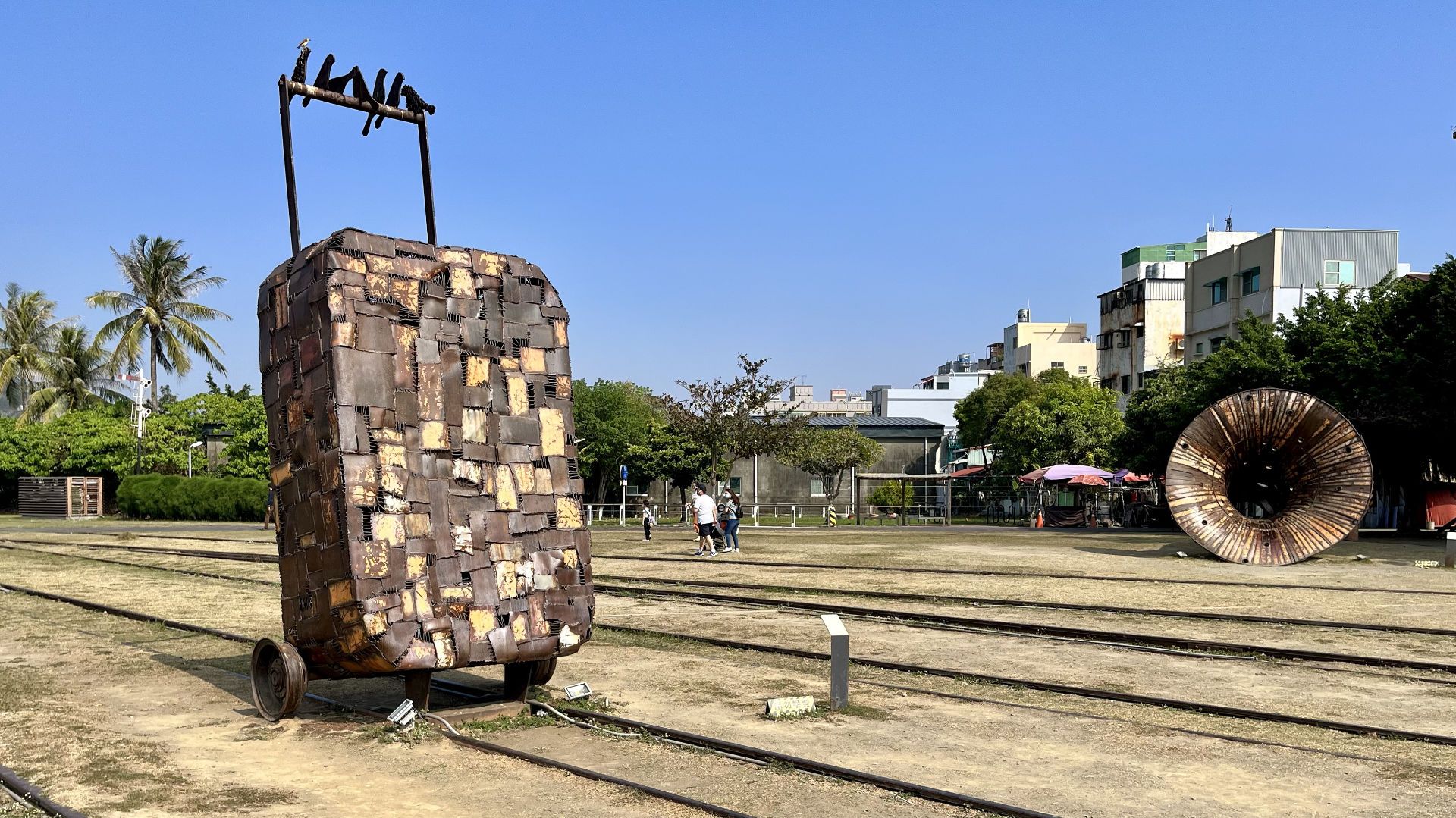 A sculpture of a suitcase on wheels, approximately 5 meters tall, with a giant horn sculpture in the background.