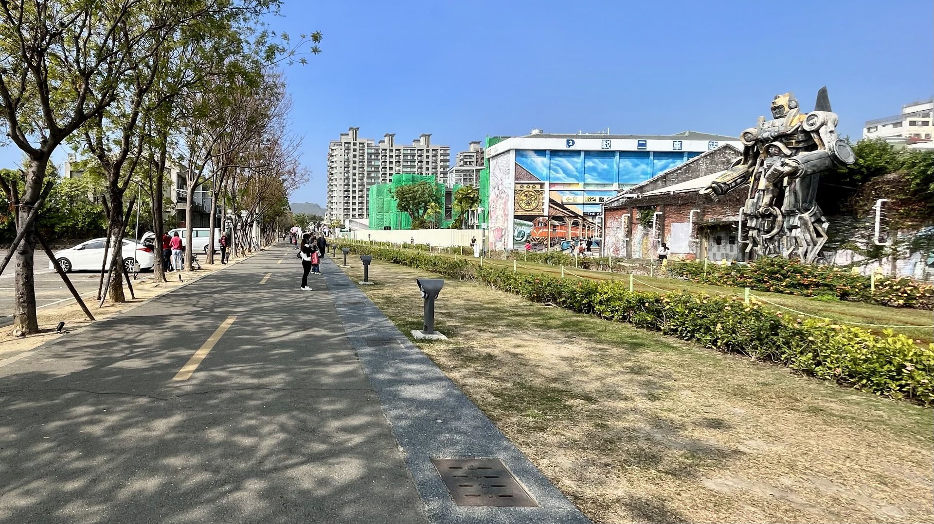 A long straight path under trees, with a sculpture of a Transformer-like robot watching from the other side of the tram tracks.