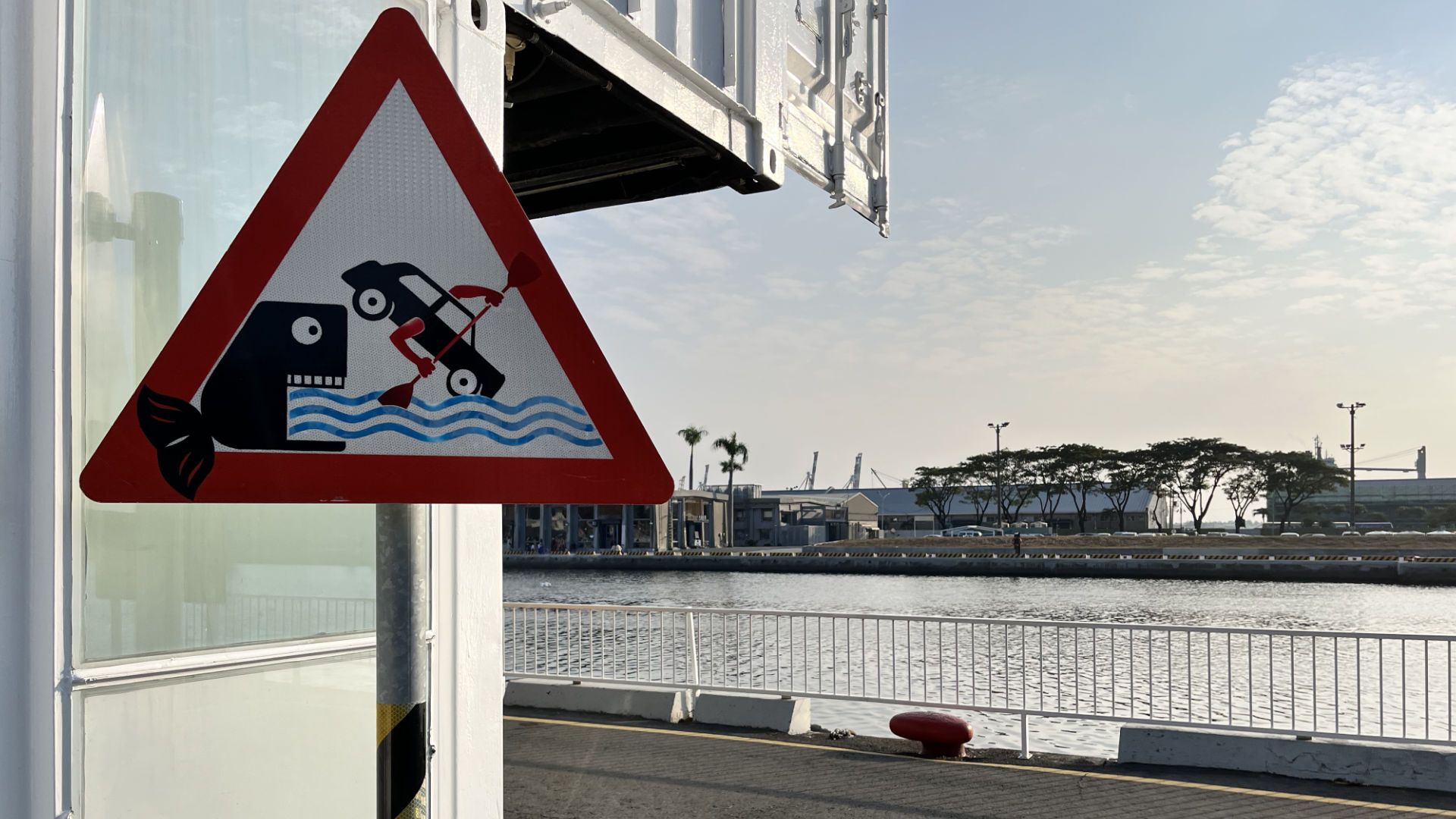 A triangular road sign depicting a large fish biting a car that is being rowed along a body of water.