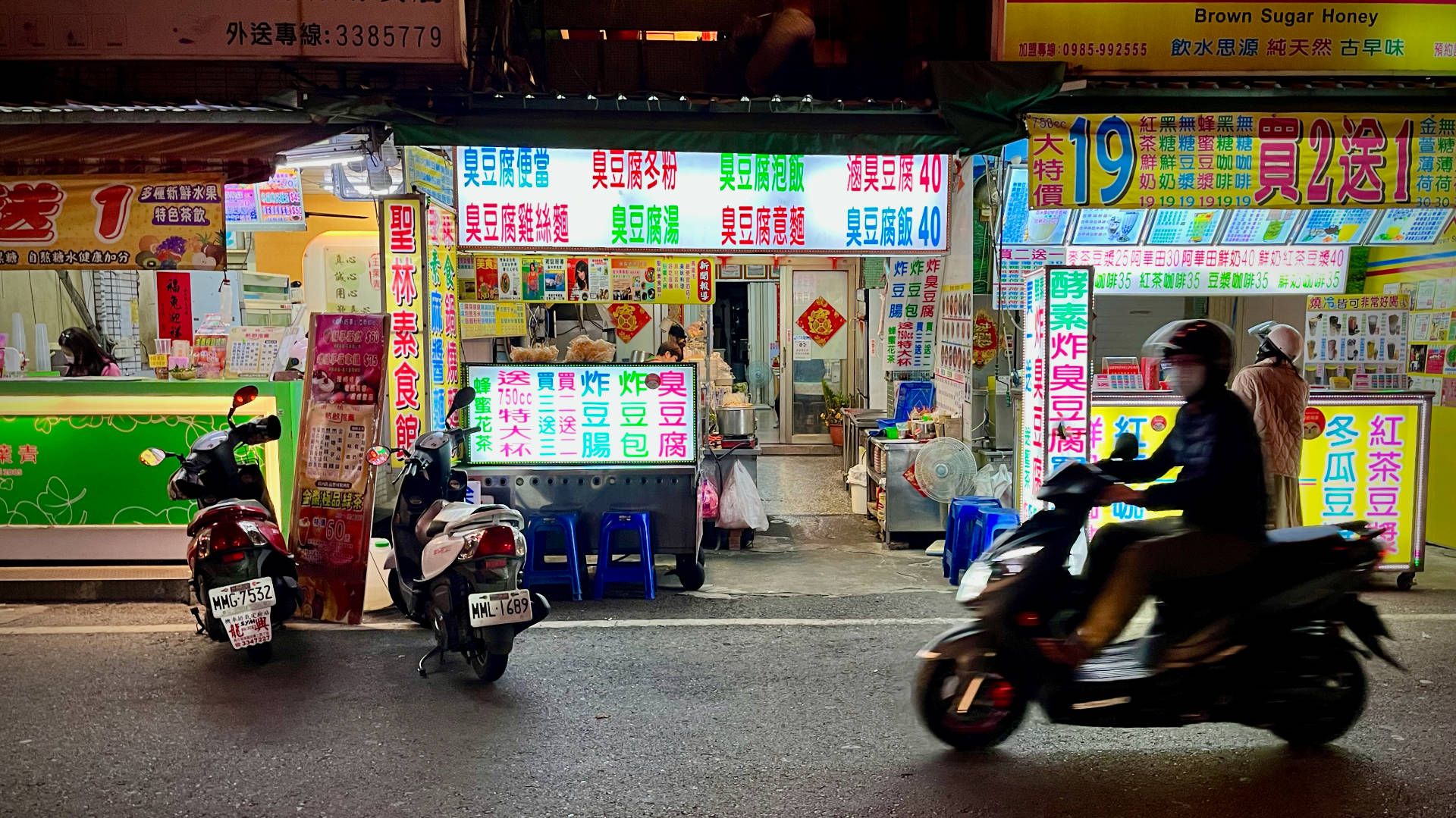 Photo of a restaurant with bright signage written in Traditional Chinese. Two scooters are parked outside, and a third scooter is being driven past.
