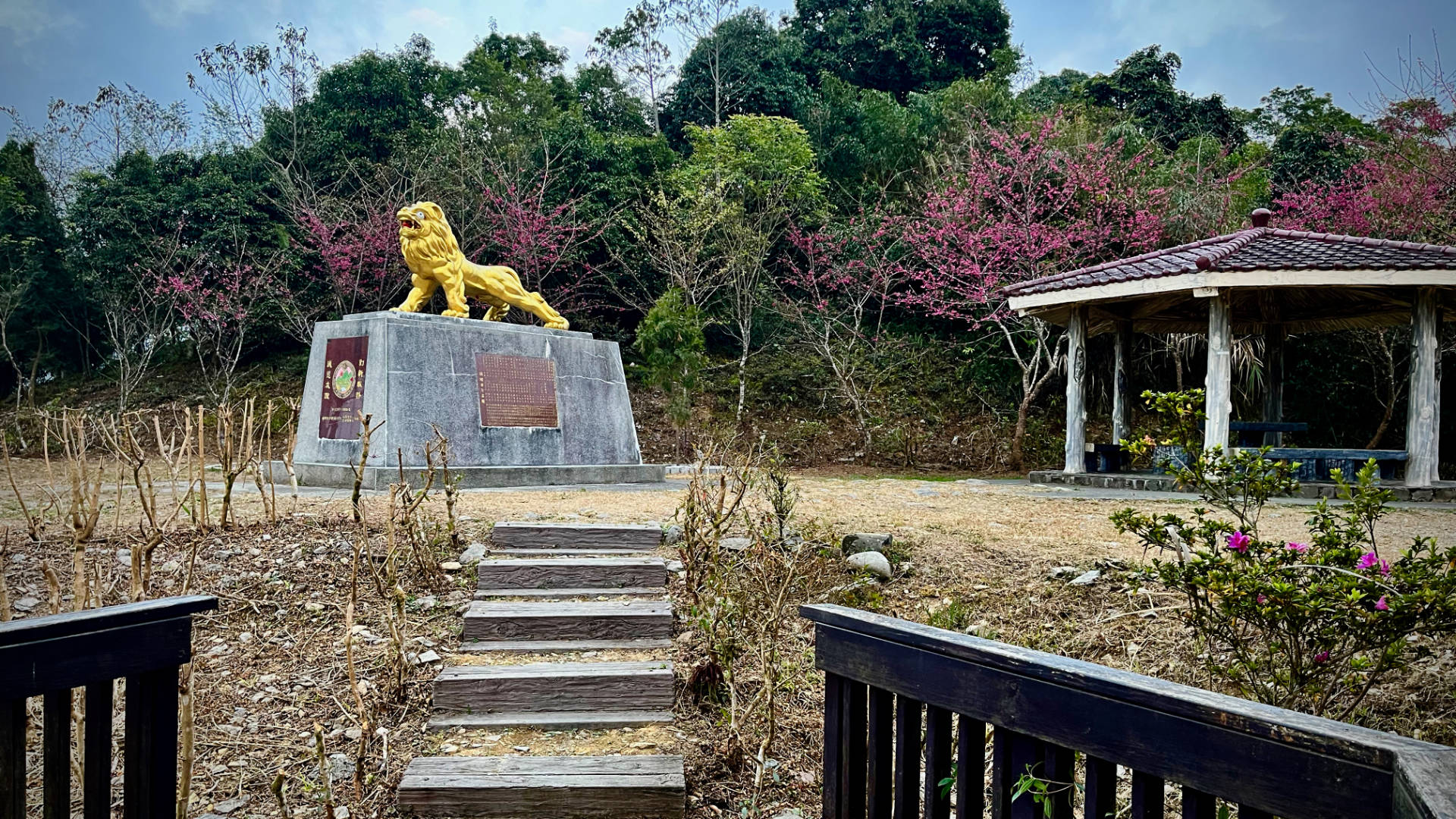 Steps leading up to a lion statue, about 1 meter tall, atop a 1.5 meter tall gray concrete base, with a gazebo behind it.