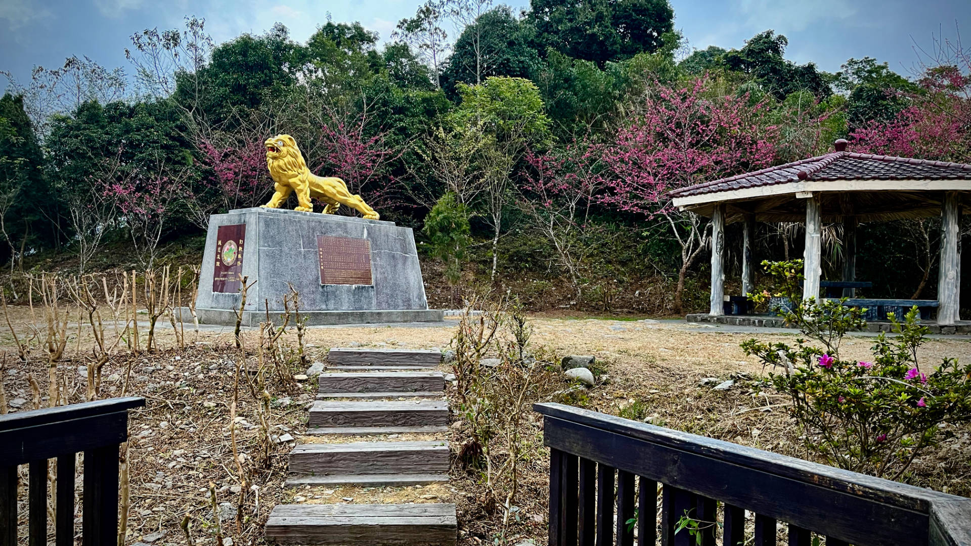 Steps leading up to a lion statue, about 1 meter tall, atop a 1.5 meter tall gray concrete base, with a gazebo behind it.