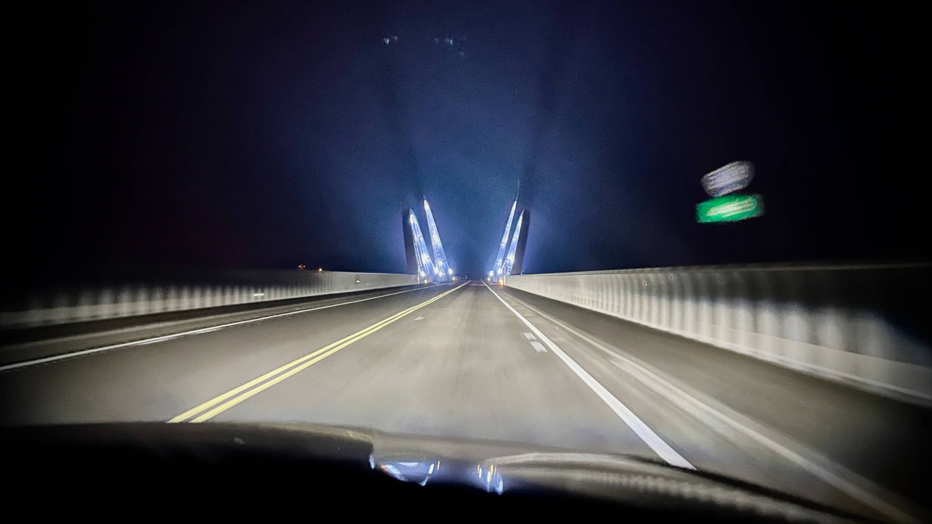 Photo taken from the front seat of a moving car, showing a long and empty two-lane bridge, with illuminated arches in the distance.