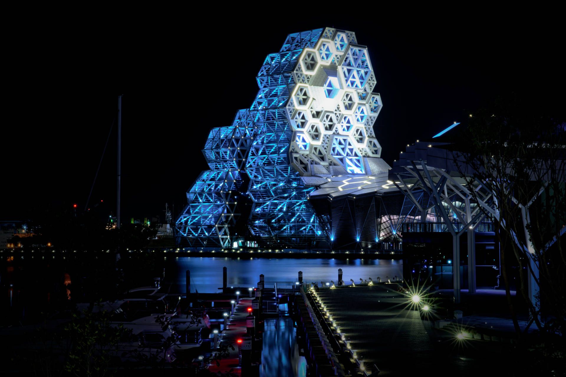 The Kaohsiung Music Center illuminated in turquoise, beyond a marina and the Argo Yacht Club building.