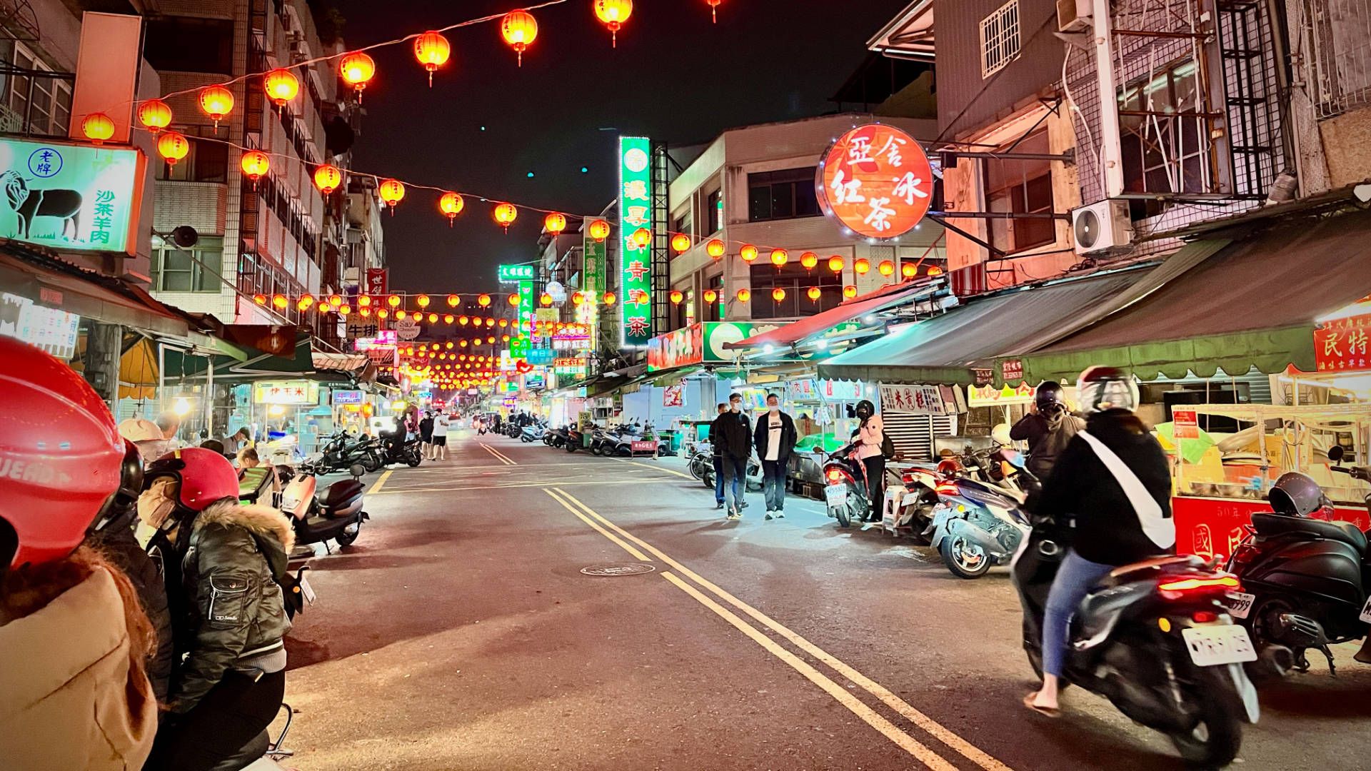 A wide-angle shot of the night market on Fuxing 2nd Road. People are walking or riding scooters along the street, and red lanterns hang overheat.