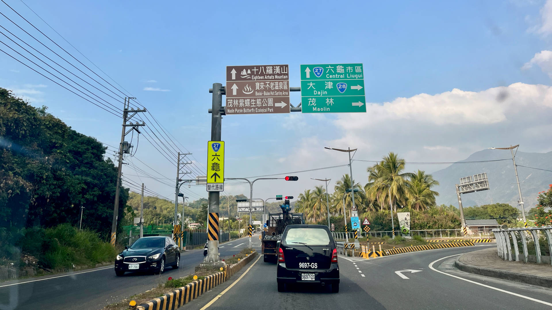 A photo taken from the front seat of a car stopped at traffic lights. Signposts above the intersection list local tourist attractions. Mountains are visible in the distance.