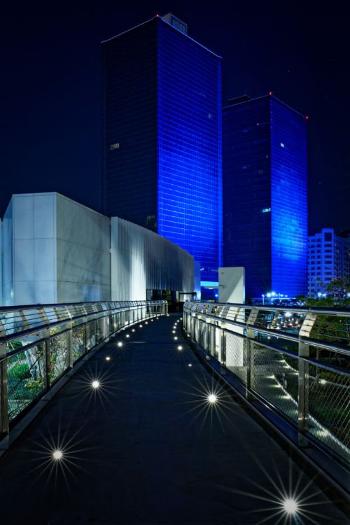 Two glass office towers illuminated in blue, viewed from an elevated walkway at Glory Pier.