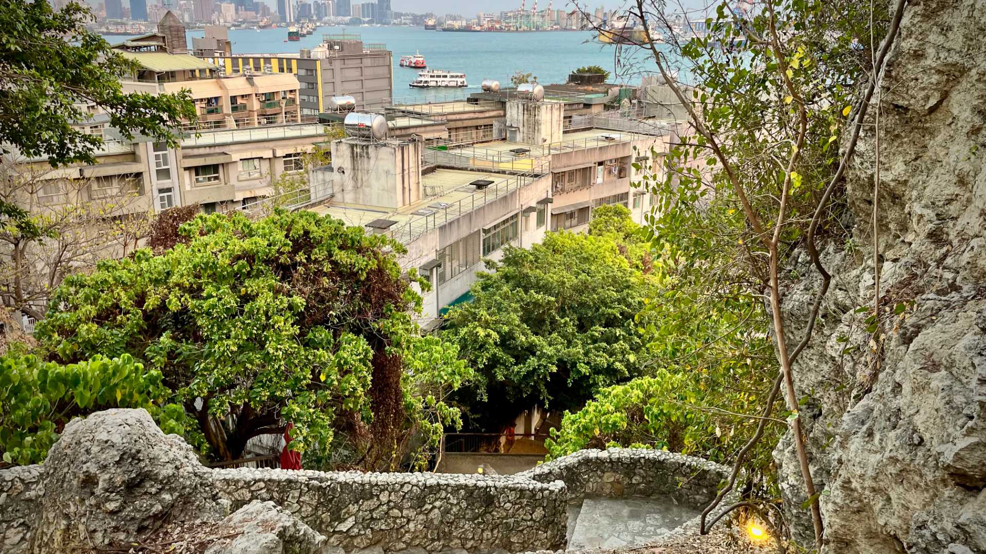 Looking downhill over the stone steps and apartments towards Kaohsiung Harbor. Two scooter ferries are visible in the middle of the harbor, and cranes and skyscrapers can be seen in the far distance.