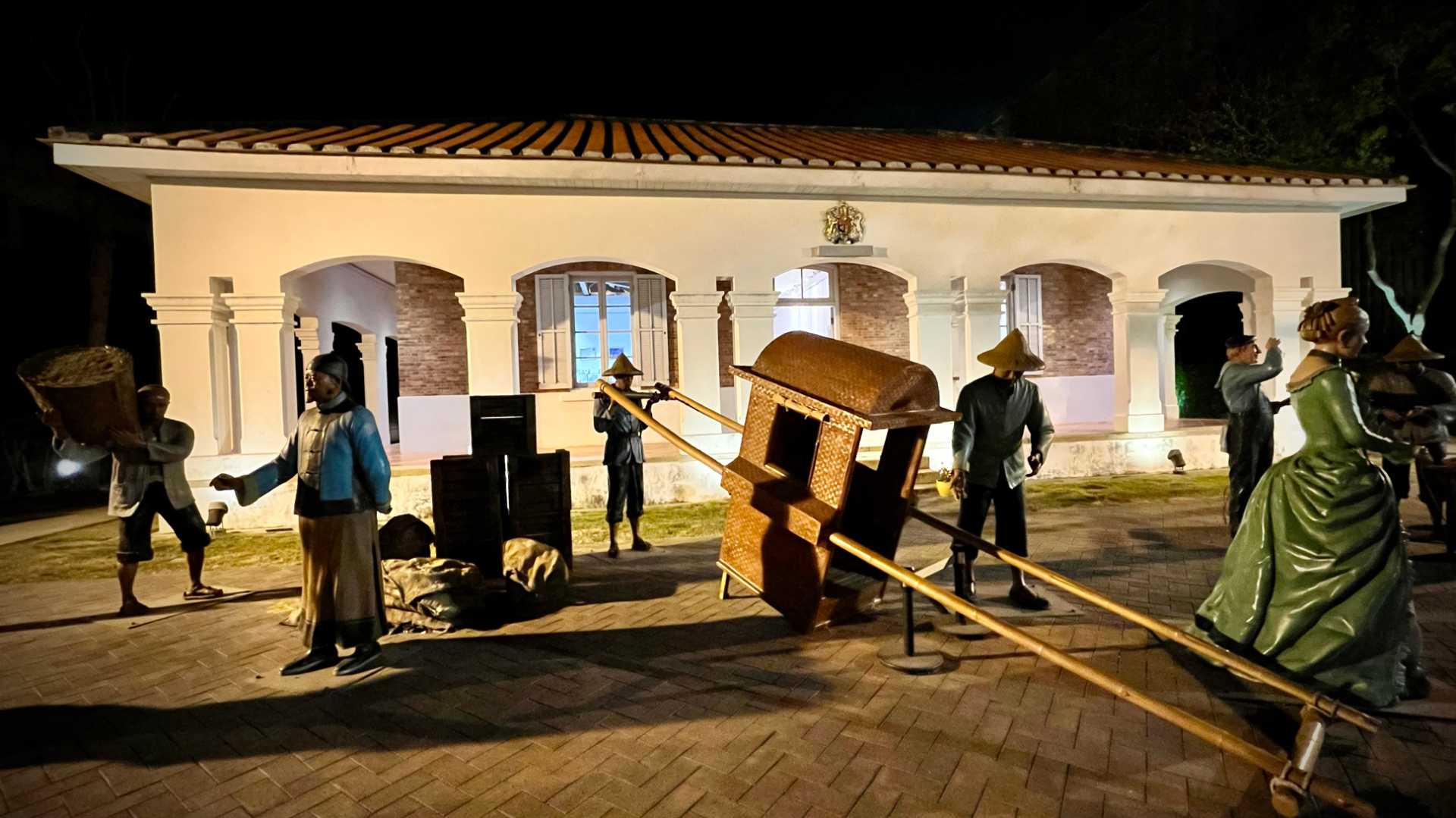 Life-size sculptures of a historic street scene outside the former Consulate Office building. Sculptures include people carrying bags and goods, and talking with each other.