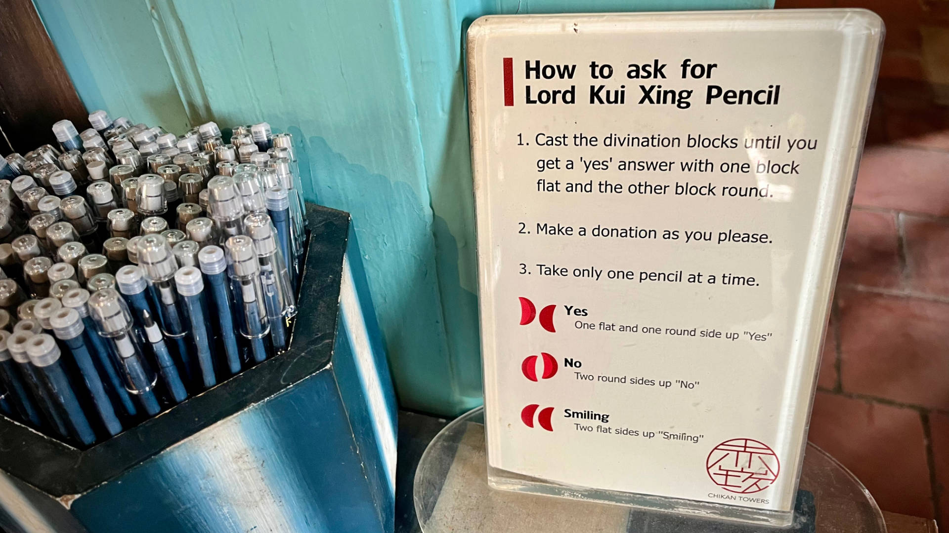 A hexagonal wooden container full of pencils, and an instruction sheet alongside it labelled “How to ask for Lord Kui Xing Pencil”. Step 1: Cast the divination blocks until you get a ‘yes’ answer with oine block flat and the other block round. Step 2: Make a donation as you please. Step 3: Take only one pencil at a time. Below the instructions, a diagram shows the combination of blocks that indicate “Yes”, “No”, or “Smiling”.