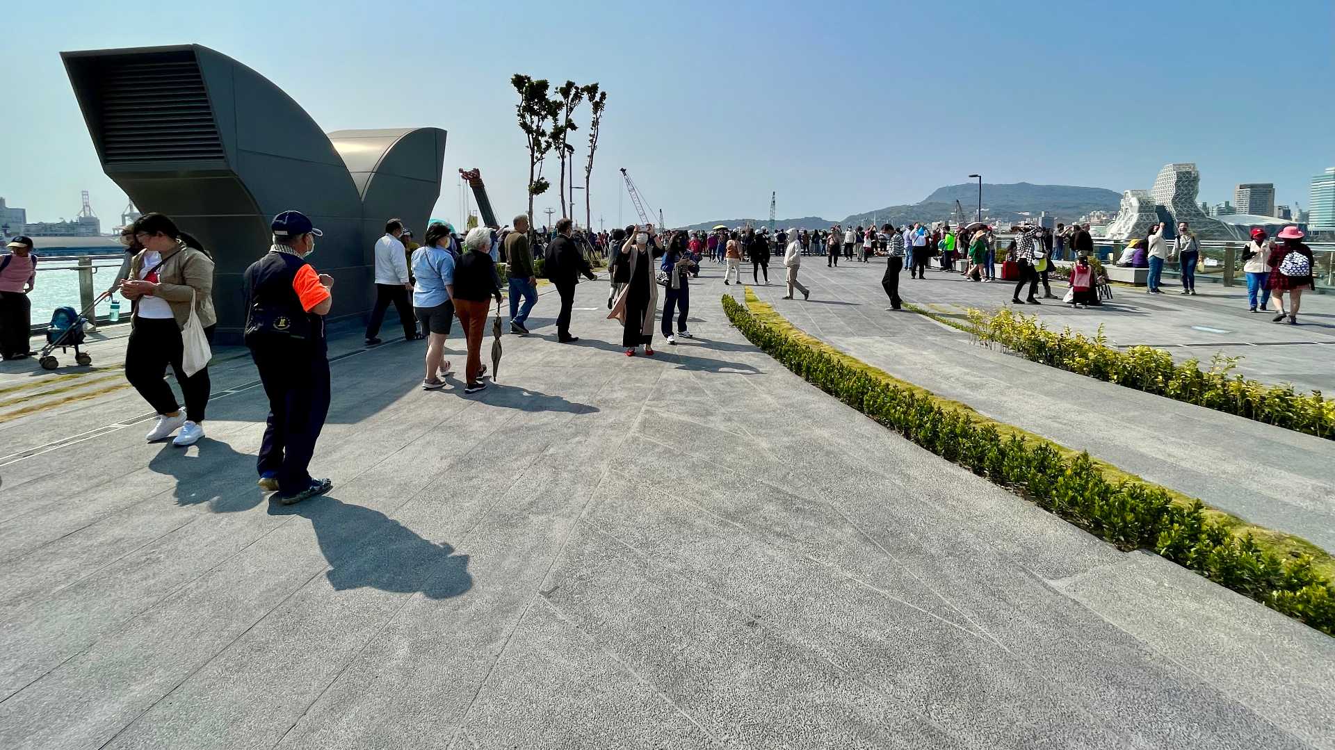 An expansive viewing deck paved in concrete with narrow curved hedge and grass sections. There are hundreds of people on the deck. A mountain and some tall buildings can be seen in the distance.