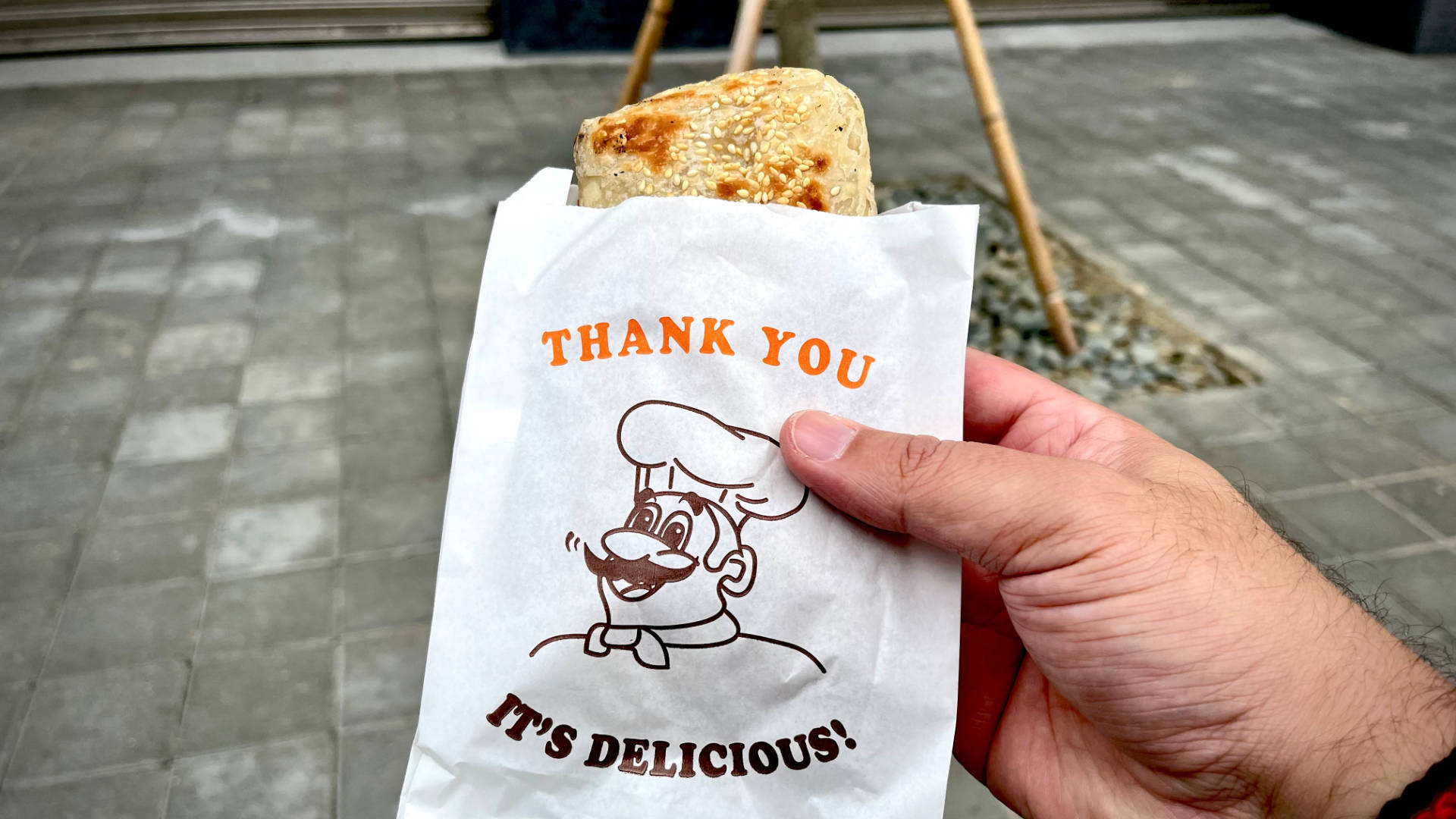 An outstretched hand holding a paper bag labelled “thank you, it’s delicious”. A pastry snack is protruding from the bag.