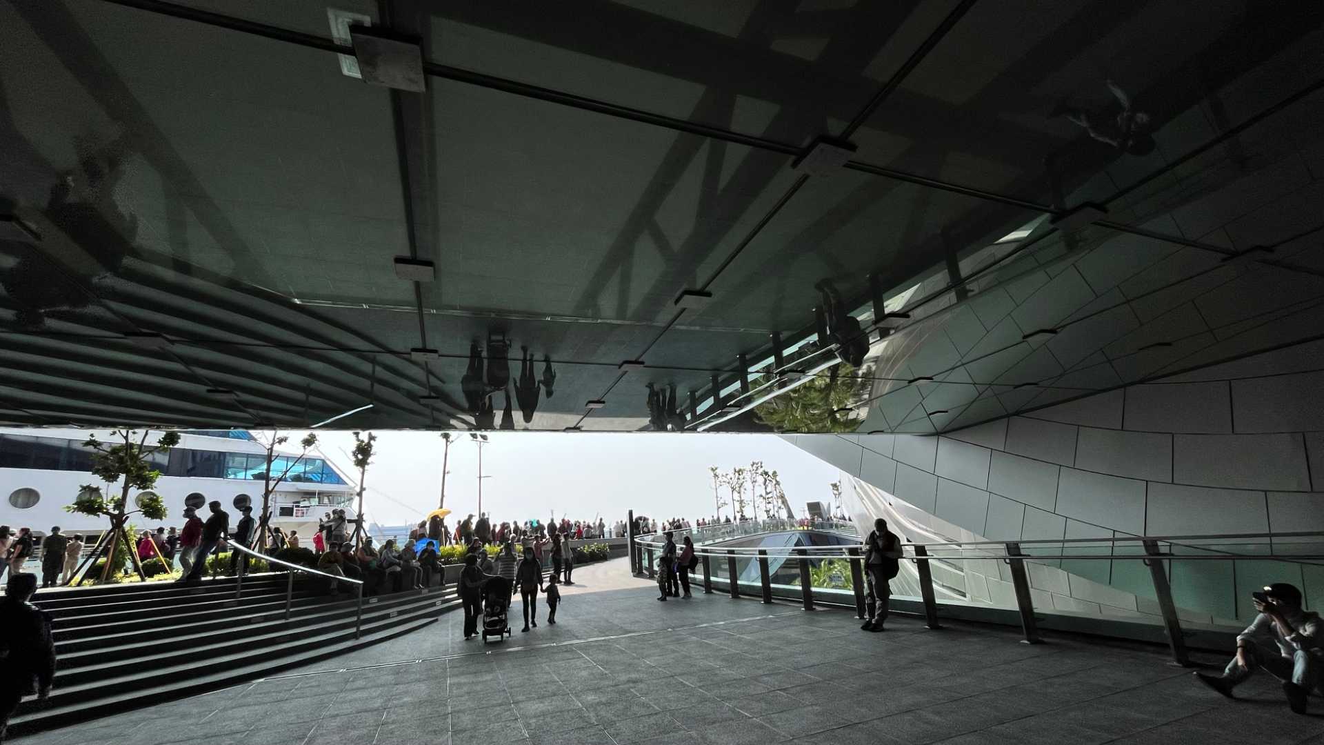 A mirrored ceiling projects out over the entranceway. Crowds of people are walking on the viewing deck behind, with the Nautica cruise ship docked alongside. The viewing deck is roughly level with the bridge of the cruise ship.