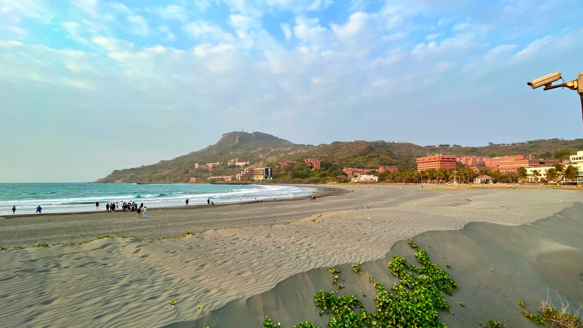 Wide-angle shot of Sizihwan Beach, with dozens of people on the beach and the buildings of National Sun Yat-sen University in the distance.