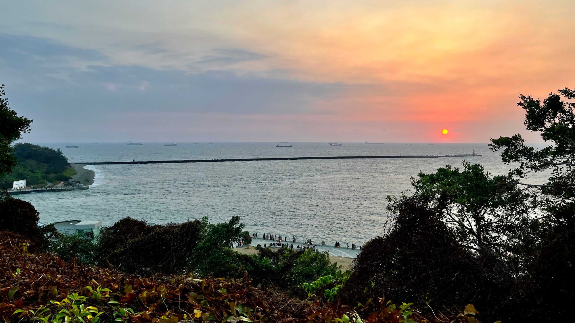 The sun setting over the Taiwan Strait. In the foreground, there are a few dozen people on the promenade watching.