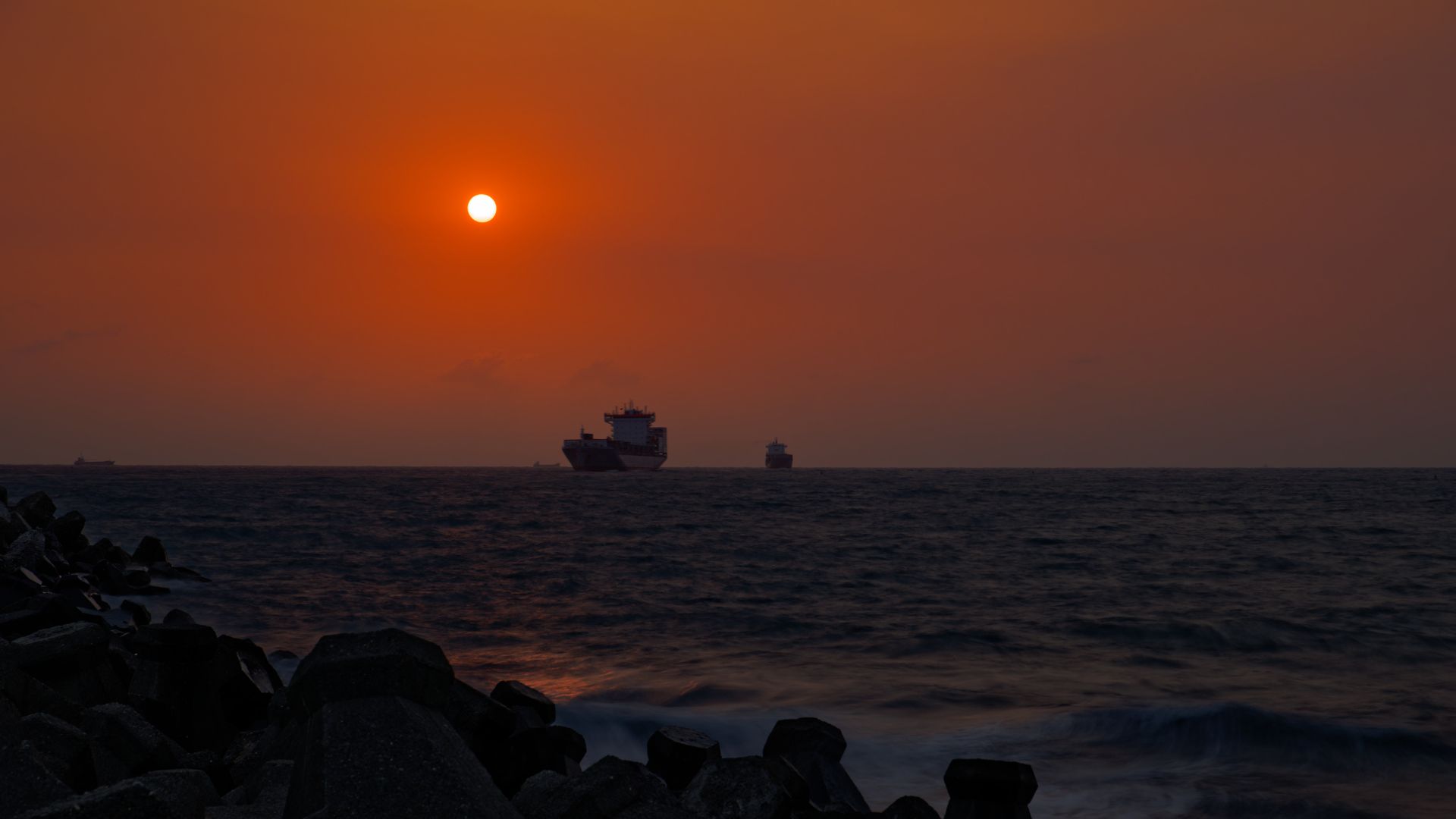 Cargo ships entering Kaohsiung Harbor in front of the setting sun. The rocks of a breakwater are visible in the foreground.