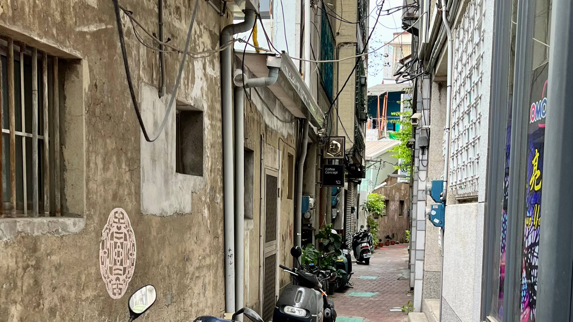 A narrow alleyway, paved in red brick with scooters parked on the left side. A mess of tangled electric cables hangs overhead.