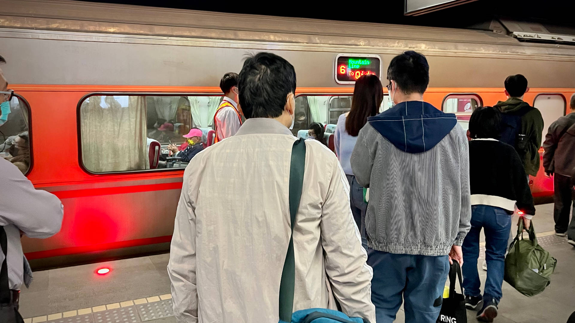Passengers queueing to board an orange-and-silver railway carriage. A sign on the carriage says “Mountain Line – 6 – To Qidu”.