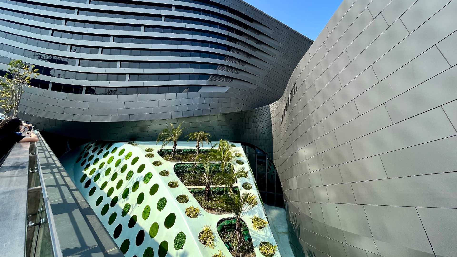 A raised white mound with palm trees planted on the top, and the sides featuring circular patches of grass, with the curvaceous metallic walls of the Kaohsiung Port Cruise Terminal behind.