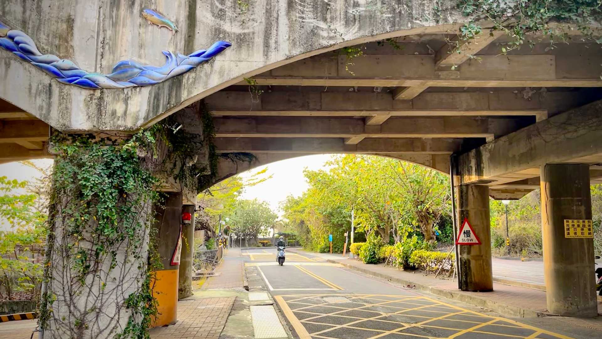 A two-lane road under a vehicle overpass, with a painted relief depicting a fish jumping from the water.