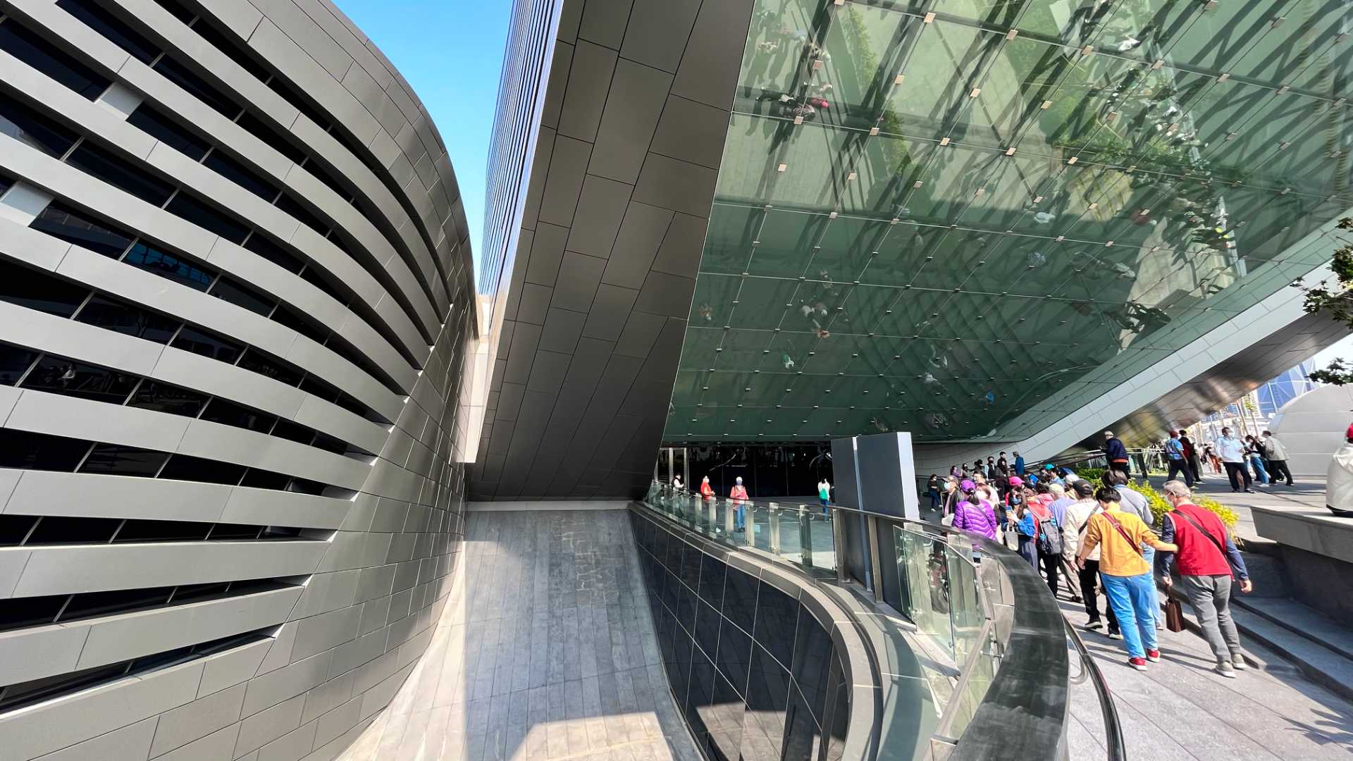View of the viewing deck entrance, showing a reflective glass ceiling projecting out over the deck. It’s a sunny day. Dozens of people are in the foreground.
