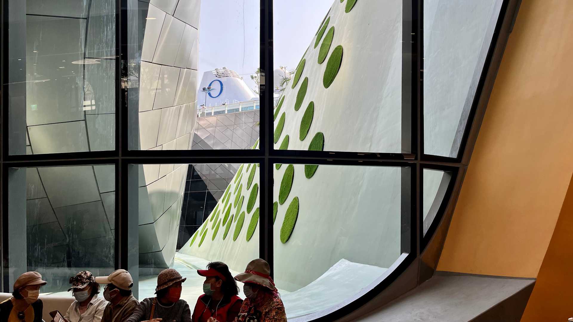 Six elderly women sit in front of a large window. They are all wearing face masks. Outside the window are the curved exterior walls, one covered in circular patches of grass, and the funnel of the Nautica cruise ship.