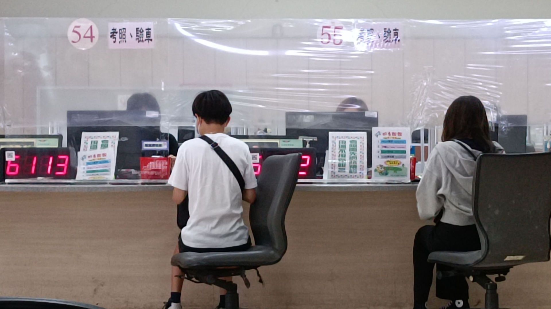 People sitting on swivel chairs at a counter, being served by administrative workers behind protective plastic screens.