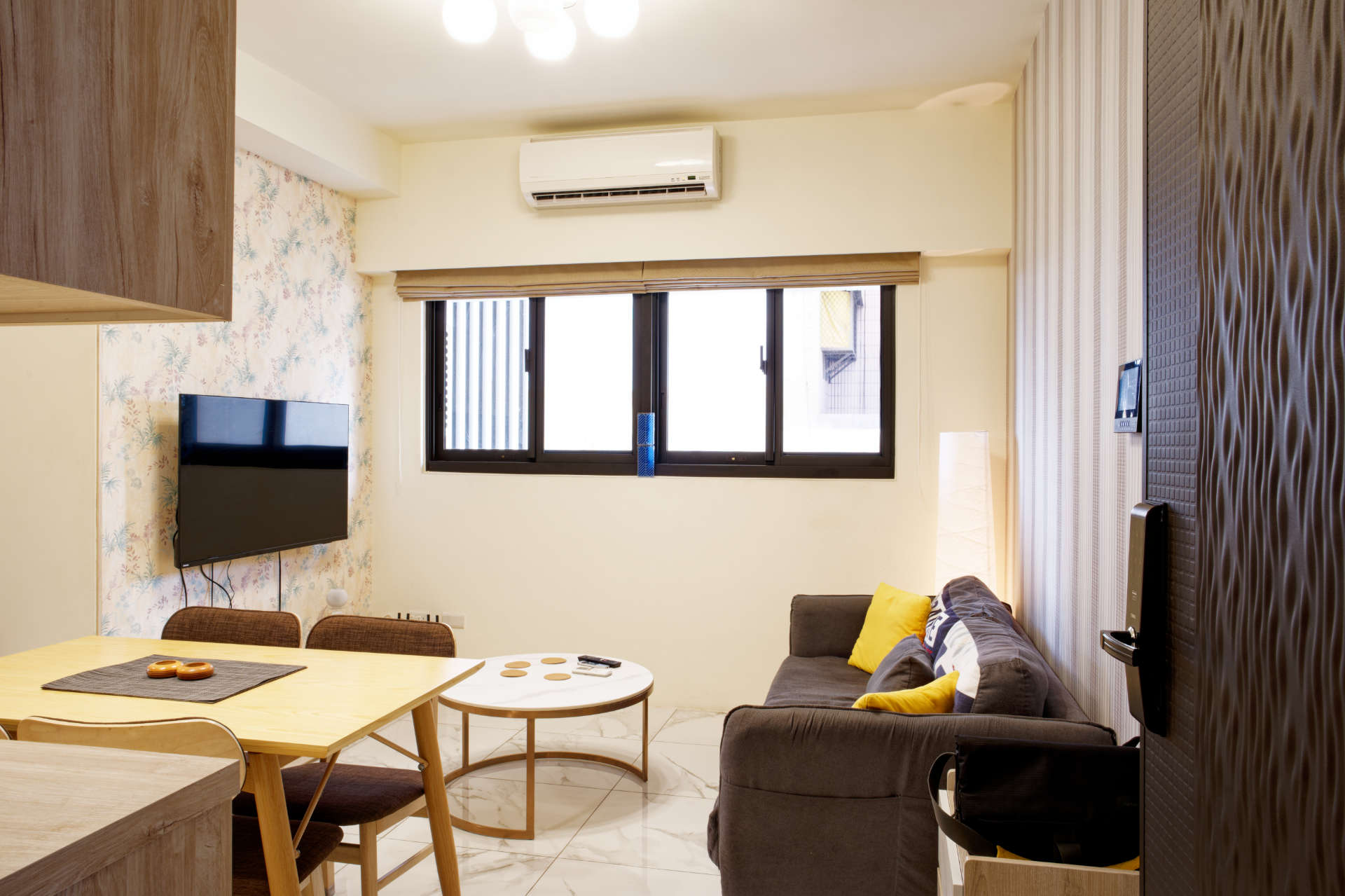 An apartment living room viewed through an open door. The room contains a two-seater couch, dining table and four chairs, a round coffee table, and wall-mounted TV.