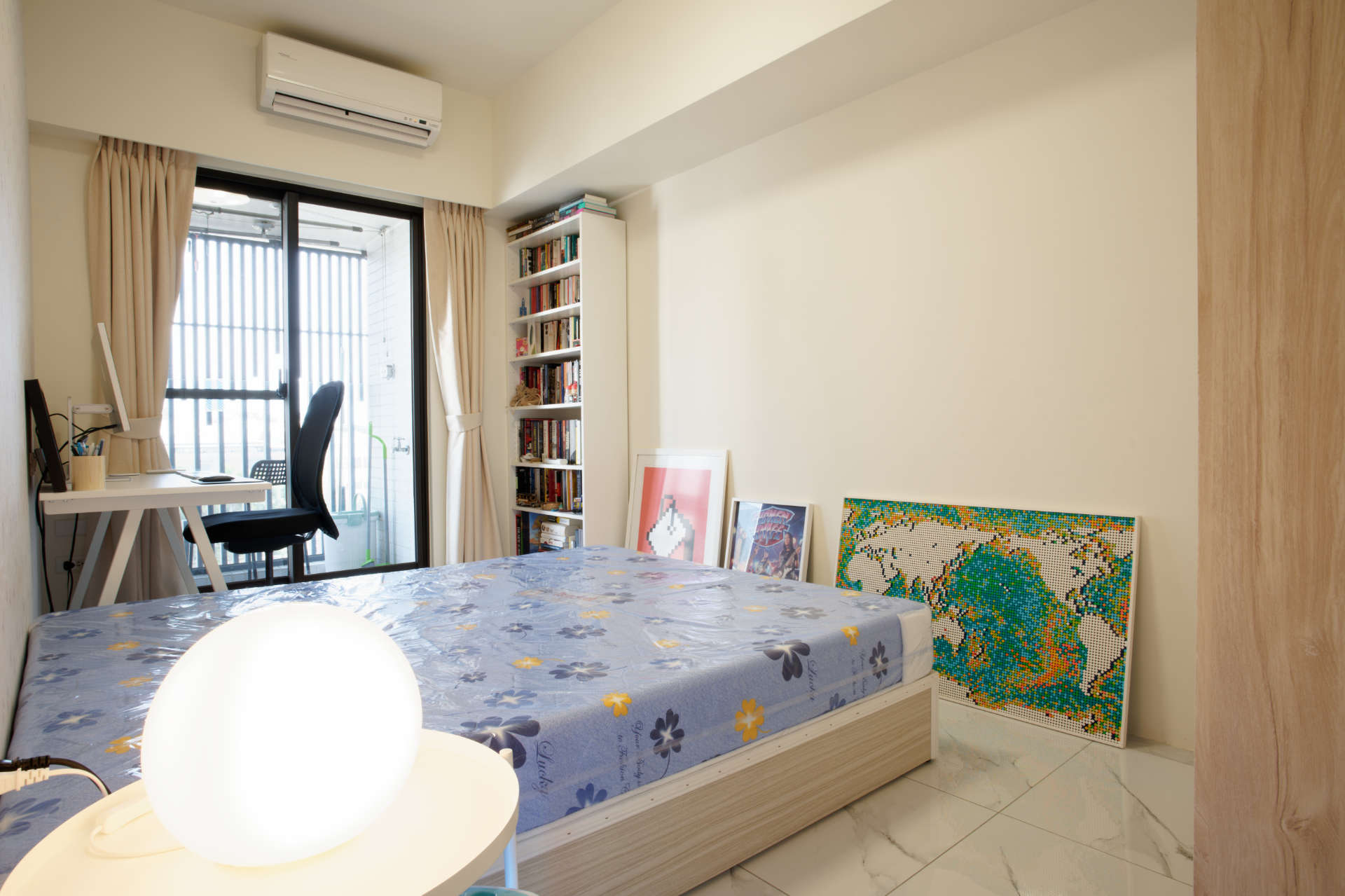 A bedroom with double bed, side table, office desk and chair, bookcase, and art leaning against the wall.