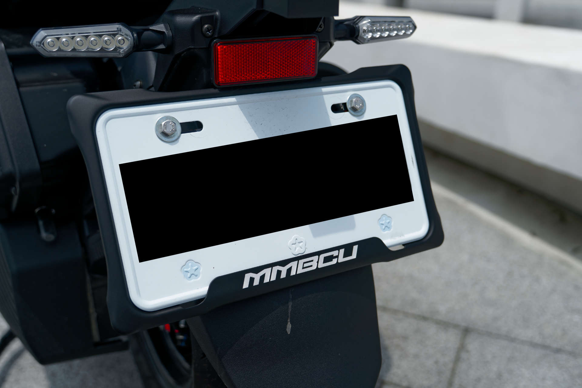 An MMBCU-branded license plate surround mounted to an SYM MMBCU scooter. The license plate number is blacked out.