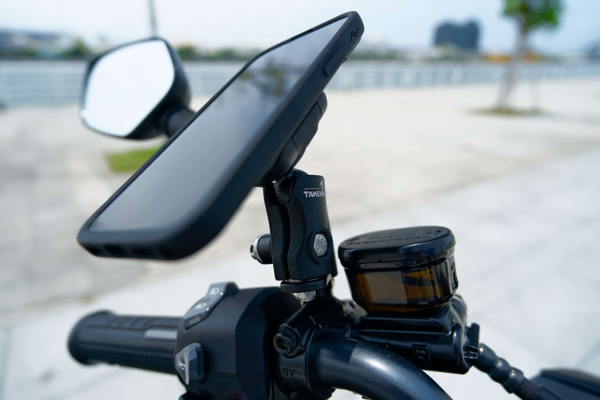 A mobile phone attached to a Peak Design ball mount adapter, held in place by a Takeway-branded base structure on the handle of a motorcycle.