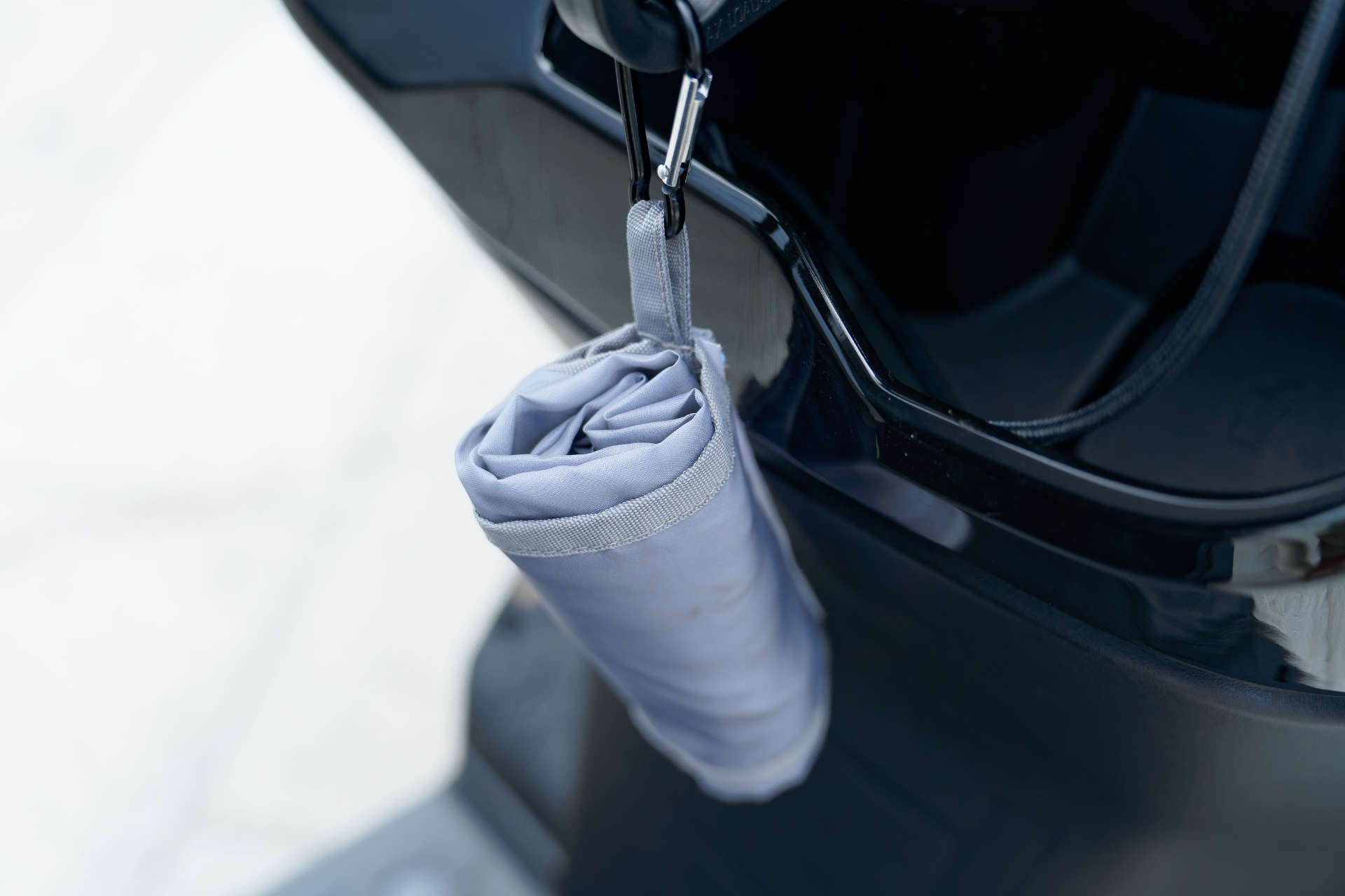 A reusable shopping back rolled up in its carry-bag, hanging from a storage hook on the front of a scooter.