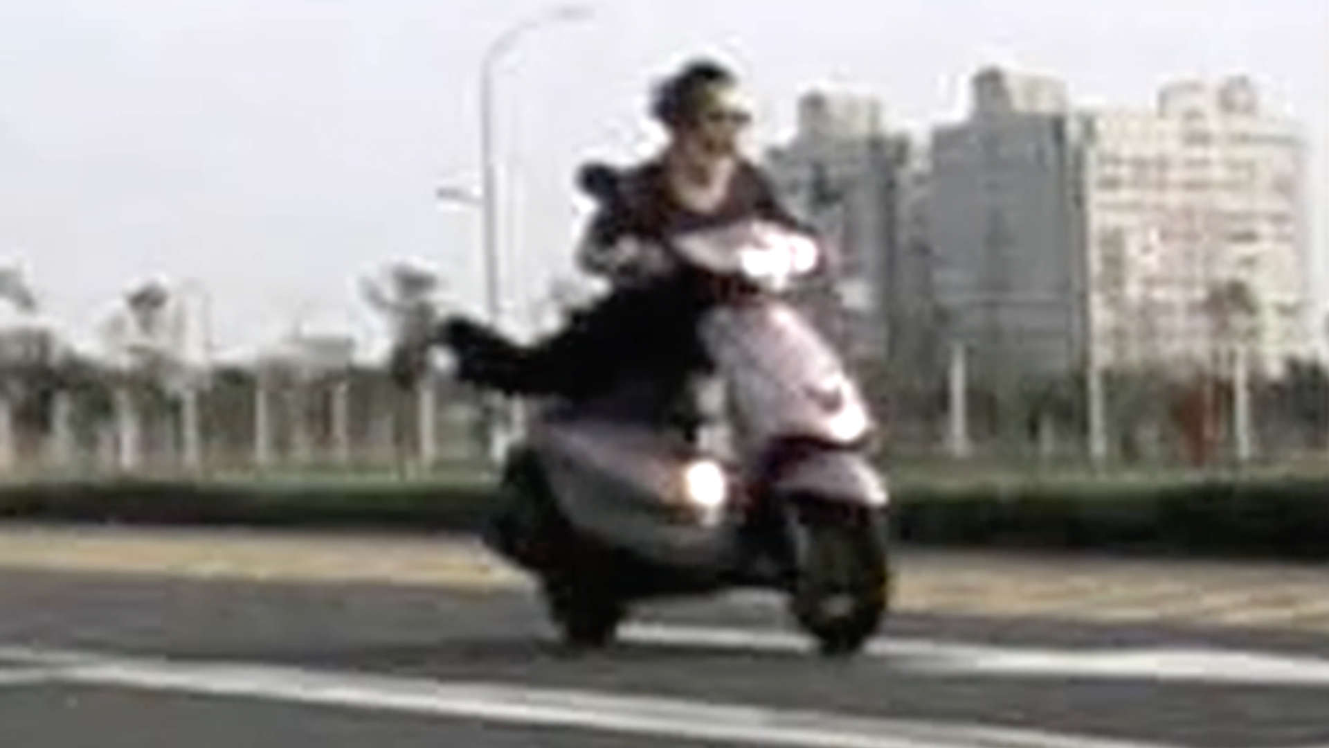 An elderly woman, without a helmet, riding a scooter face-down with feet stretched out behind her. Her hair is swept back by the wind as she rides. 