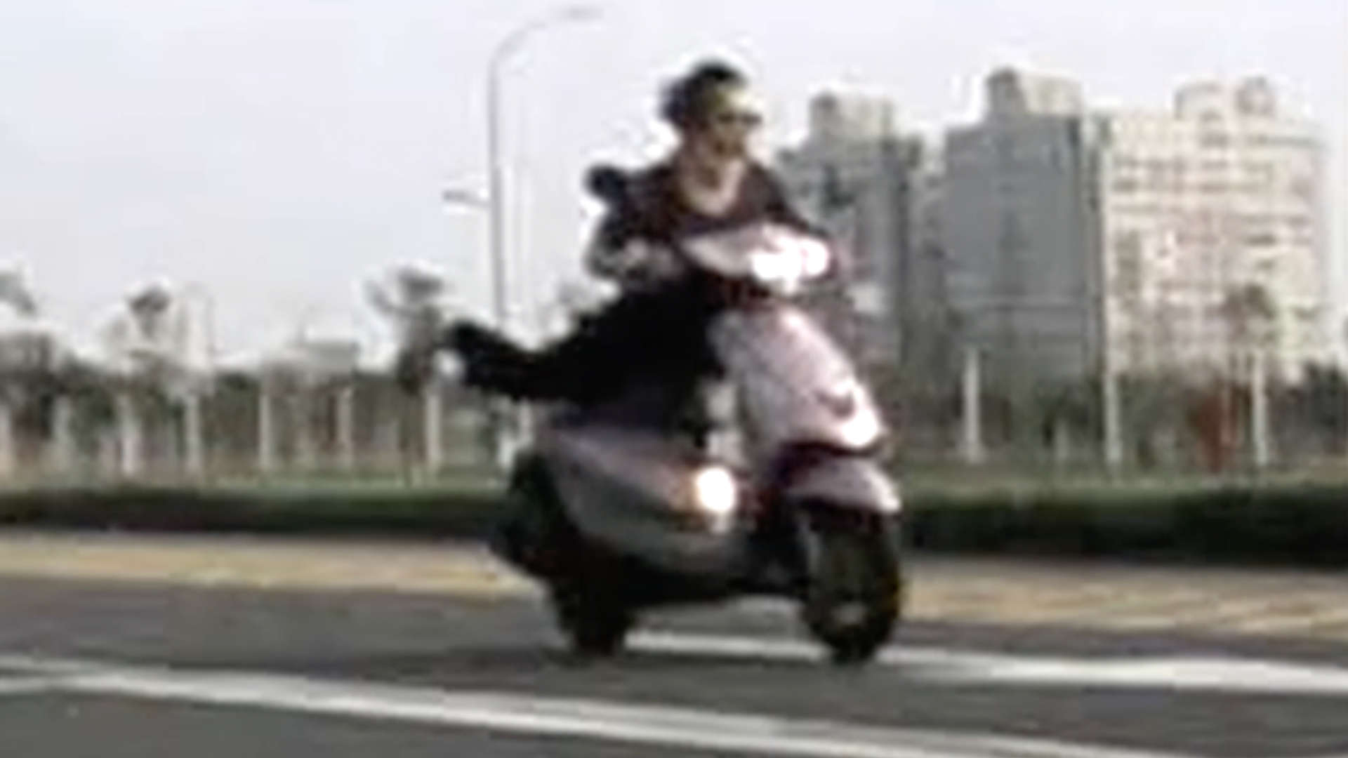 An elderly woman, without a helmet, riding a scooter face-down with feet stretched out behind her. Her hair is swept back by the wind as she rides.