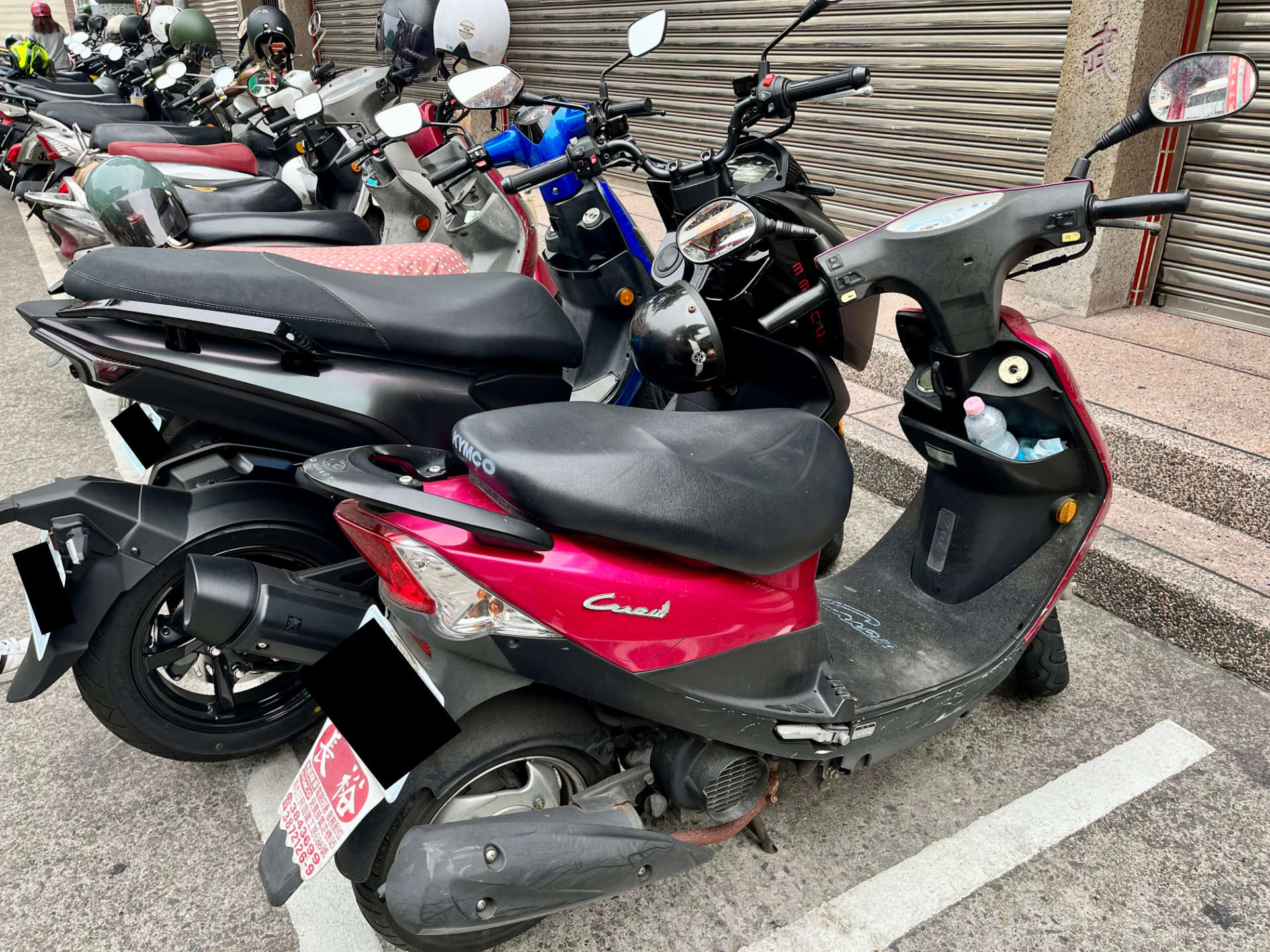 An SYM MMBCU scooter parked next to a smaller Kymco scooter, showing how the MMBCU is significantly larger.